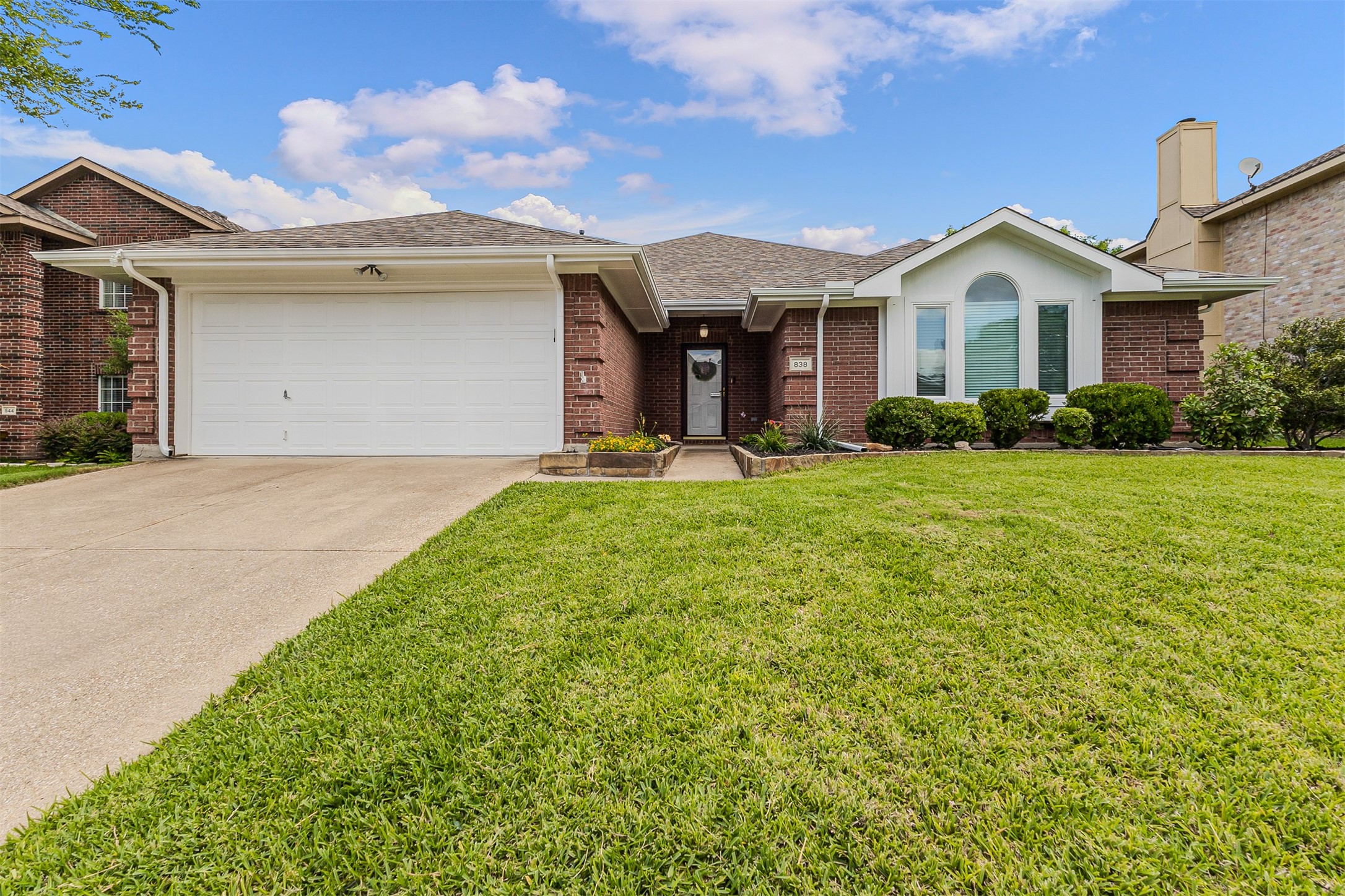 Red-brick house with a white double garage door, arched front window, and broad green lawn.