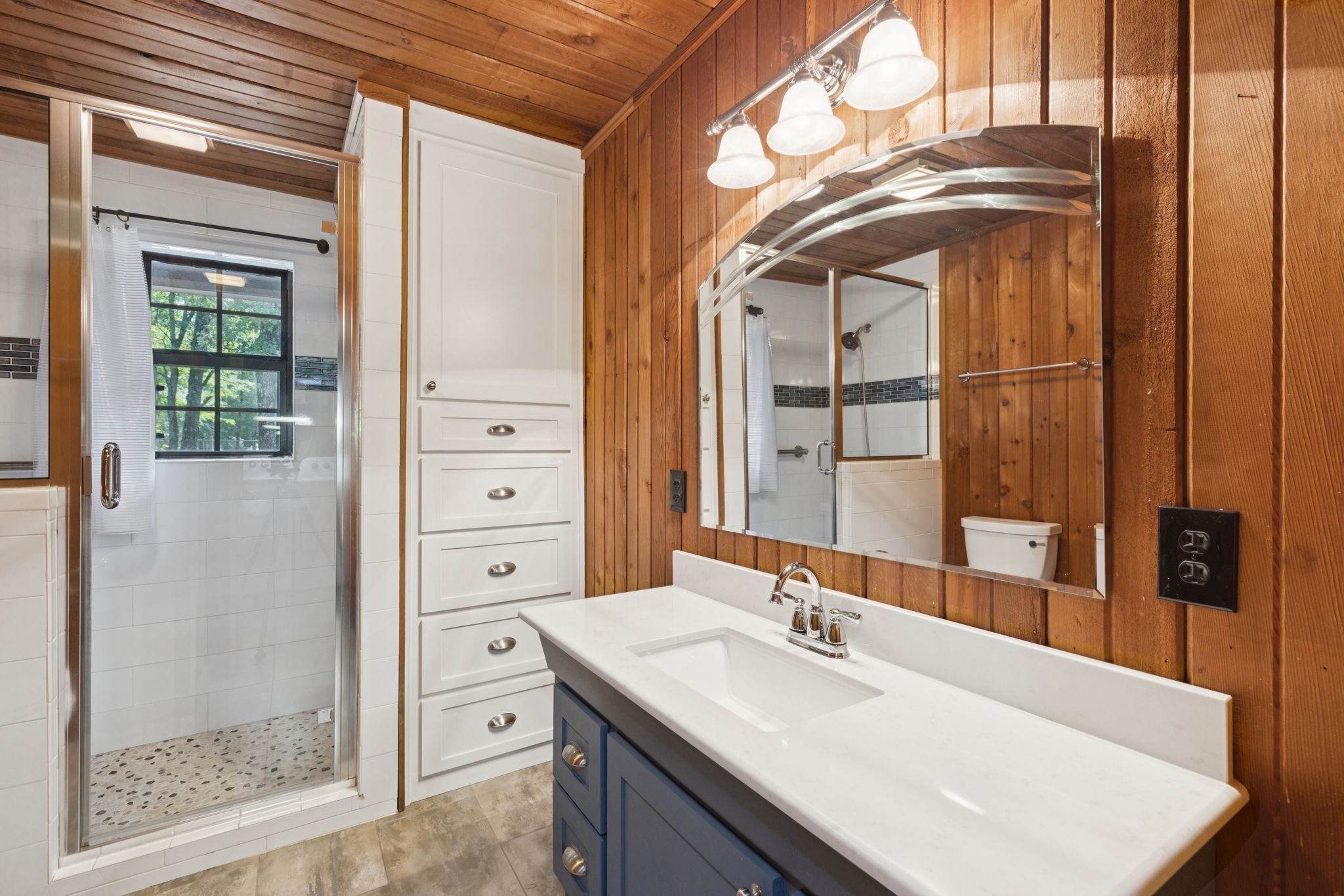 Bathroom with wood-paneled walls, a blue vanity, white countertop, and glass-enclosed shower.
