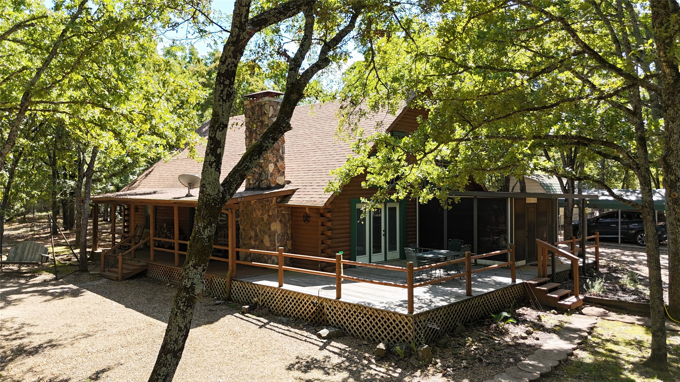 Log cabin with a stone chimney and wide deck beneath mature trees.