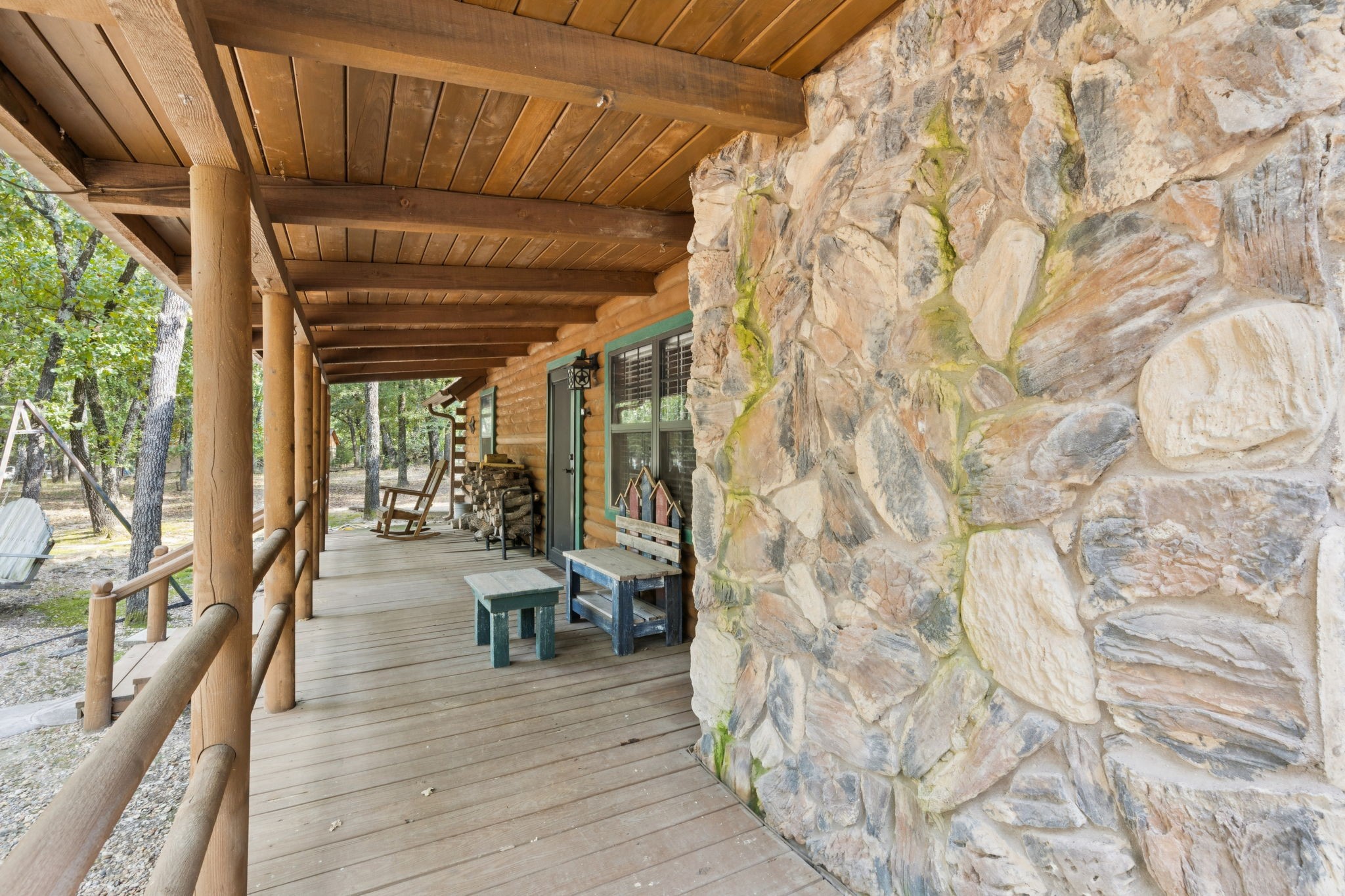 Covered cabin porch with log posts, a stone wall, rocking chair, and wooded surroundings.