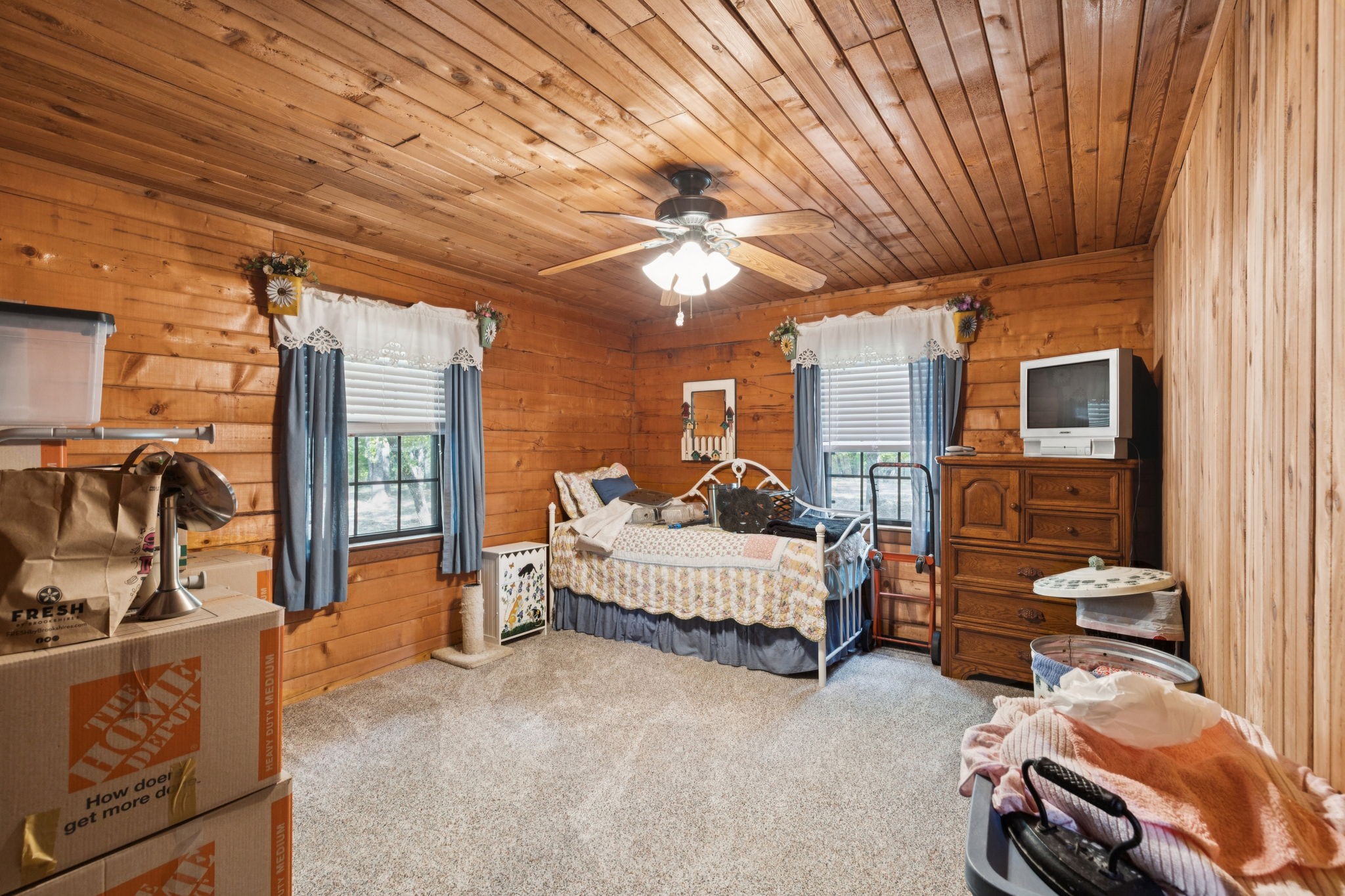 Wood-paneled bedroom with a bed, blue curtains, dresser, television, and moving boxes.