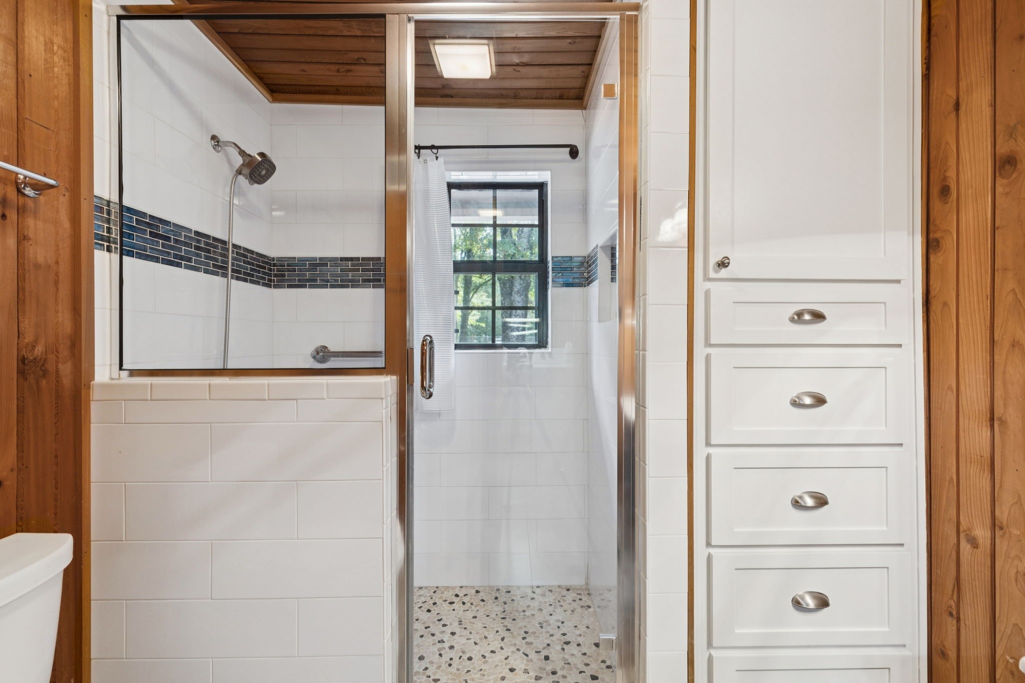 Bathroom with a glass walk-in shower, wood ceiling, tiled walls, and built-in white drawers.
