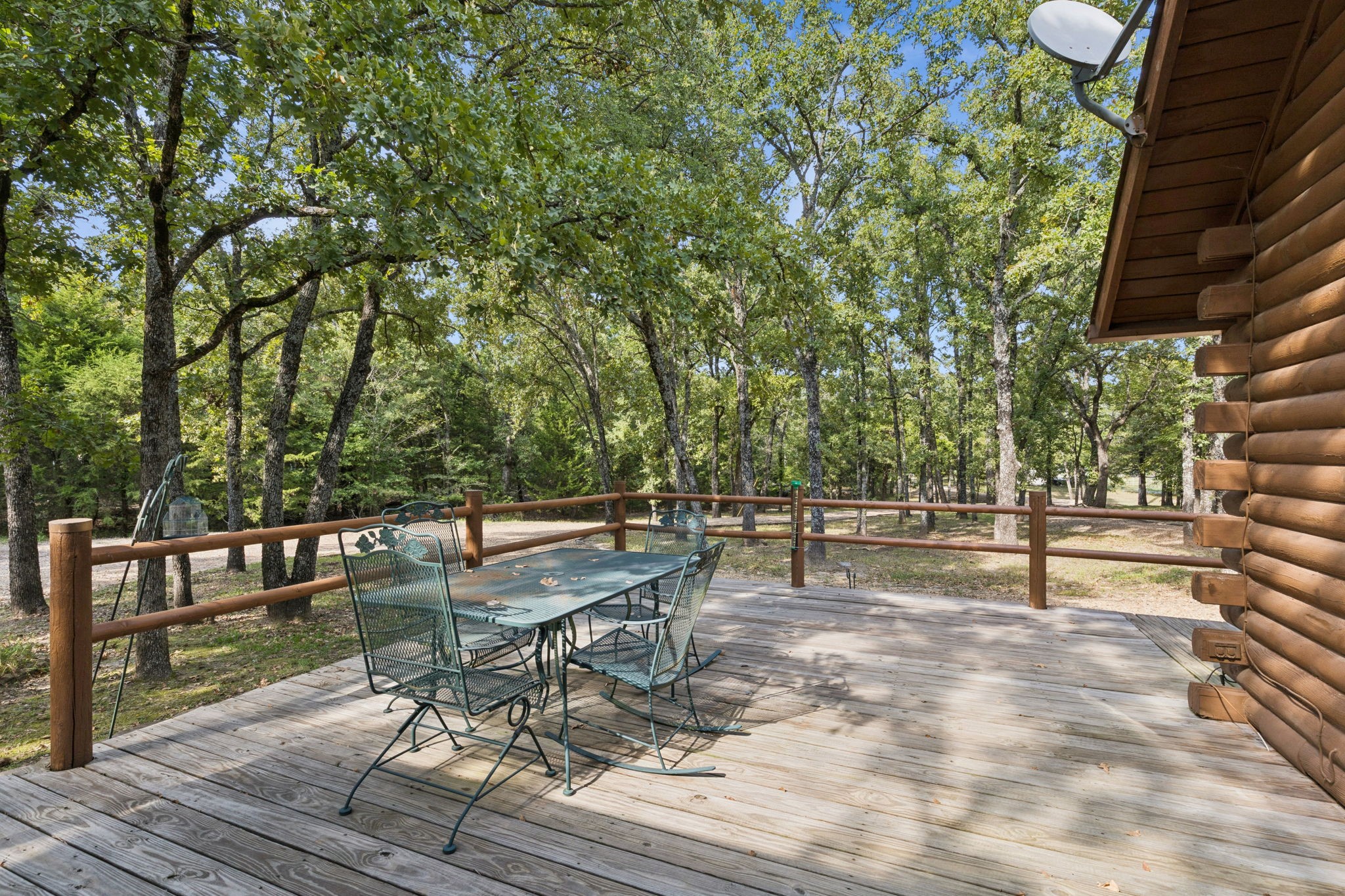 Wooden deck with patio dining set beside a log-sided home, overlooking a leafy wooded yard.