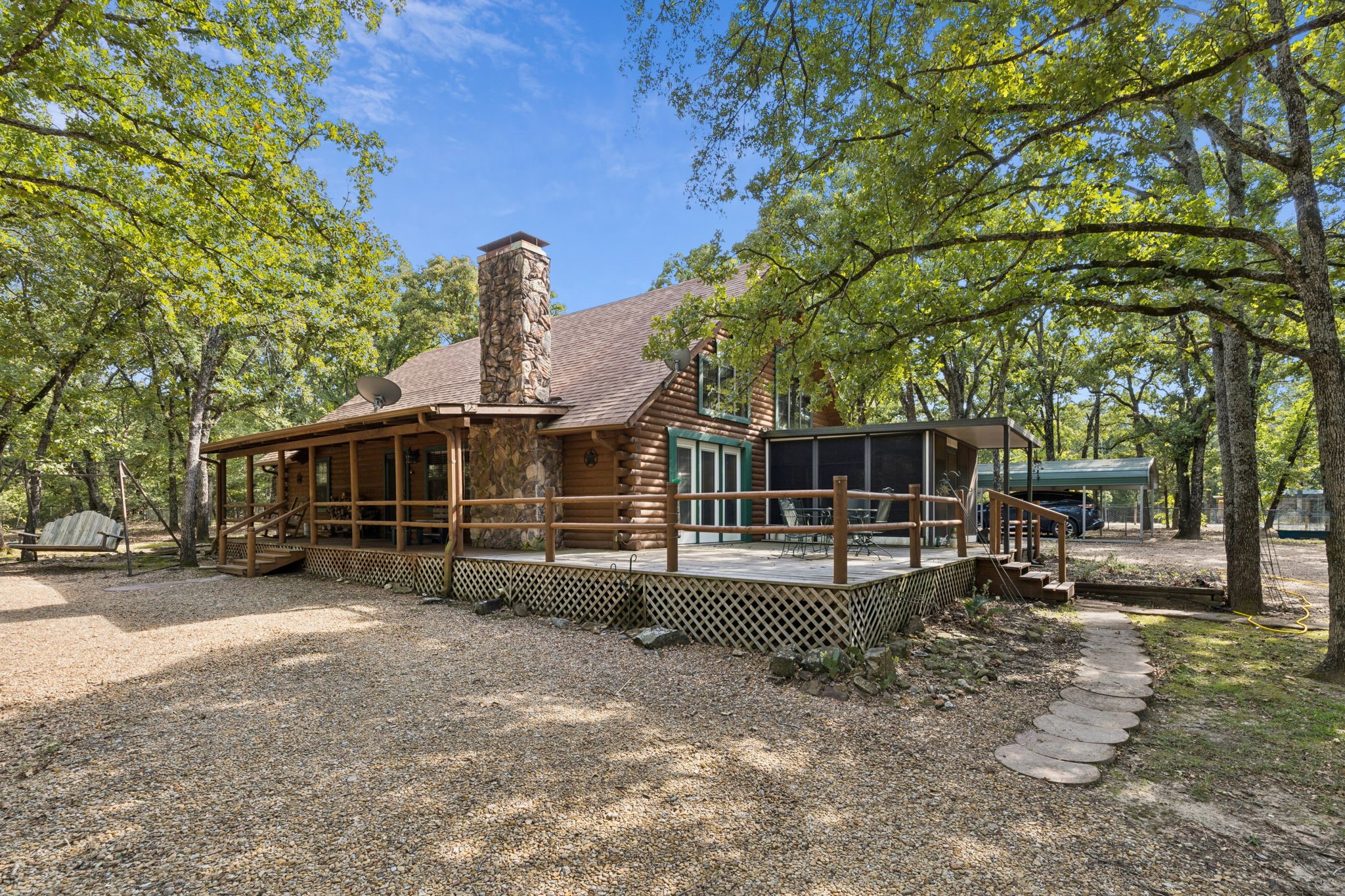 Log cabin with a stone chimney and broad deck beneath mature trees.