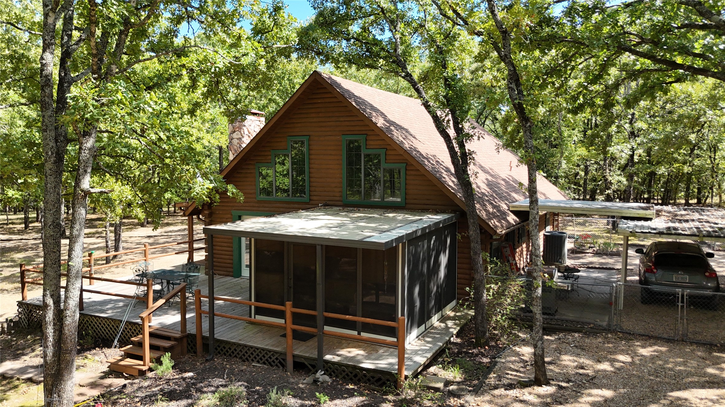 Log cabin with a covered screened porch and deck among mature trees; carport beside the house.