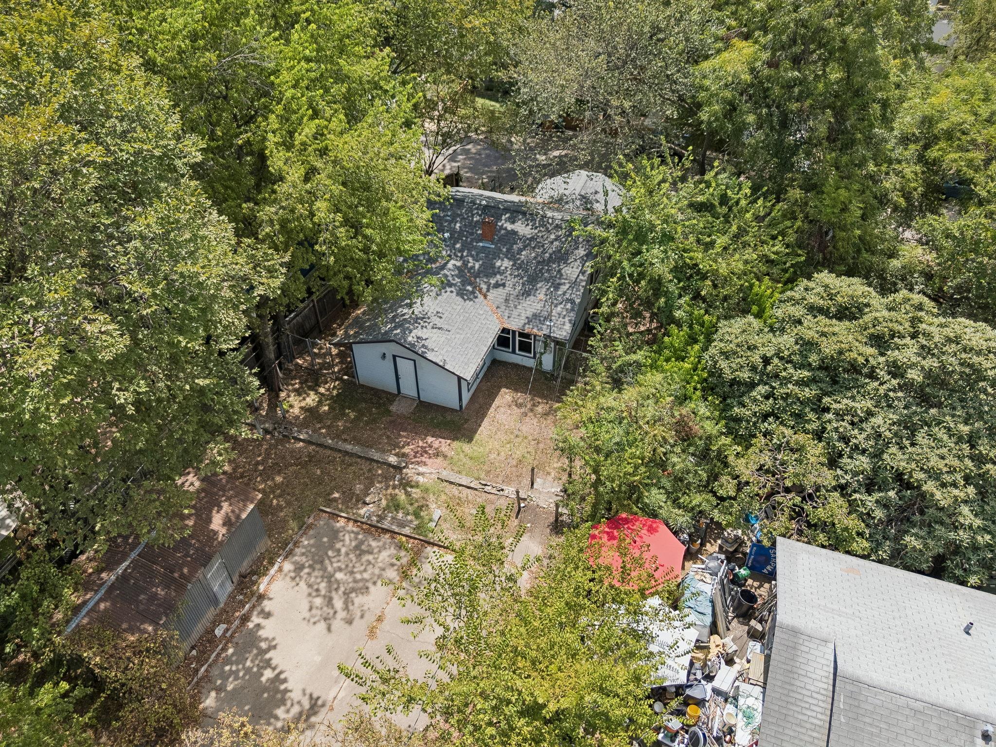 Aerial view of a small house surrounded by mature trees, with a fenced backyard and paved area.