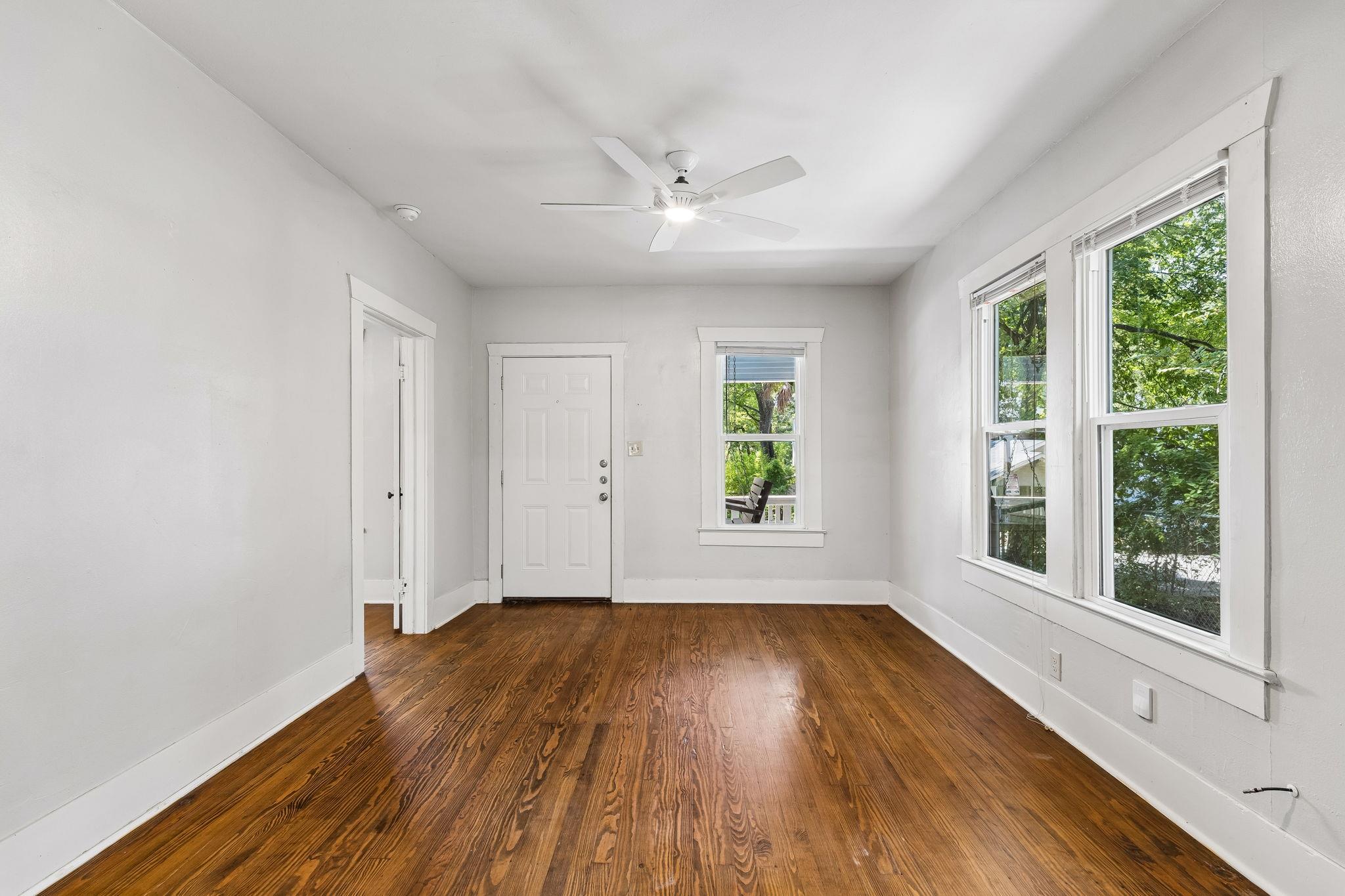 Empty room with polished hardwood floors, a ceiling fan, and large windows overlooking trees.