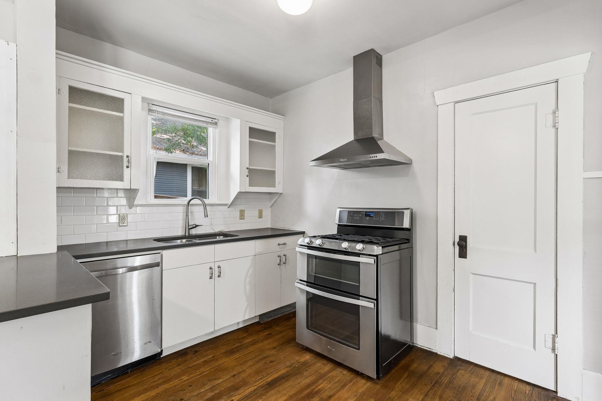 Bright white kitchen with stainless steel range and hood, dark counters, and warm wood flooring.