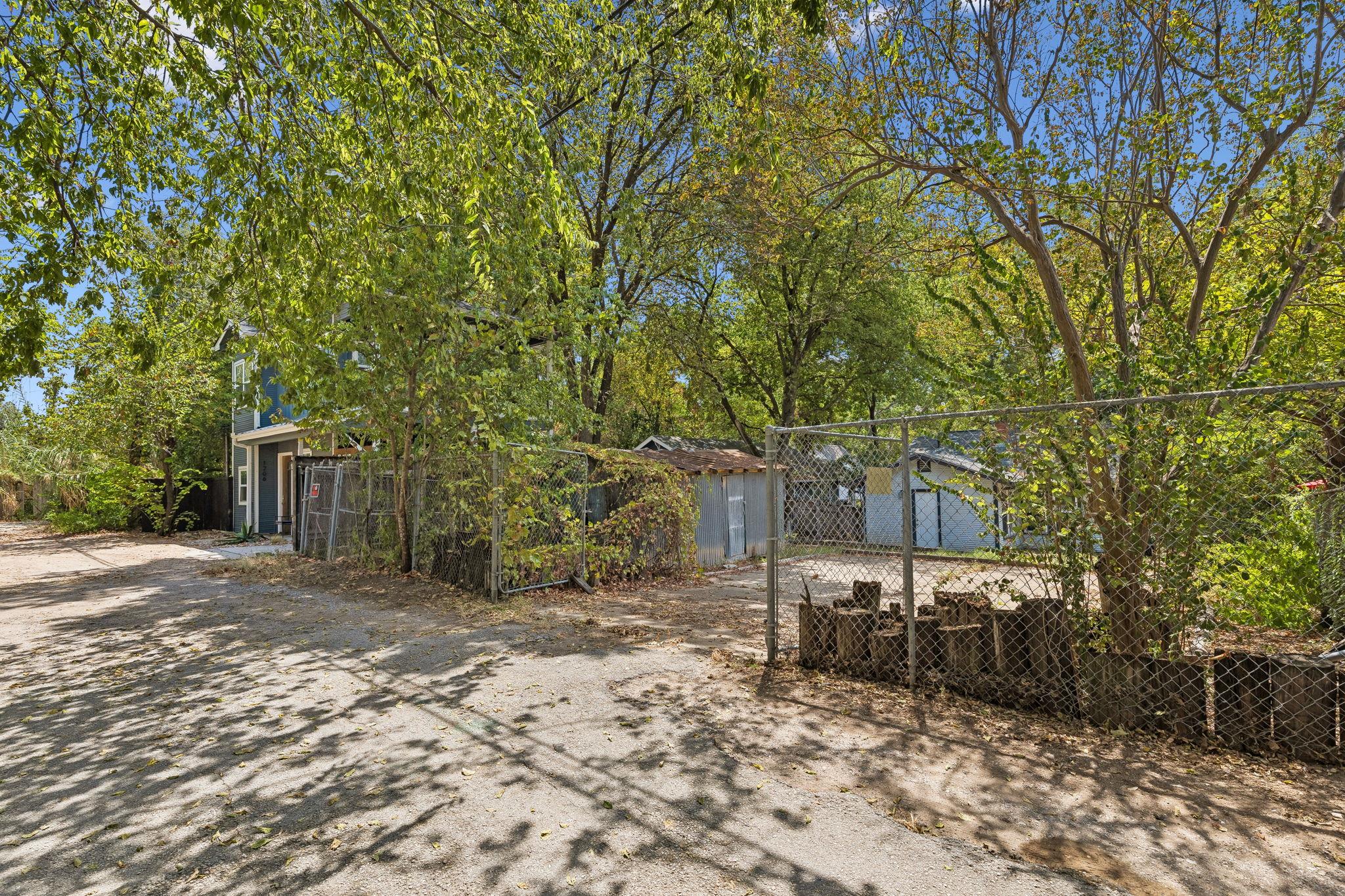 Tree-shaded property with a chain-link gate, paved driveway, and partly obscured blue house.