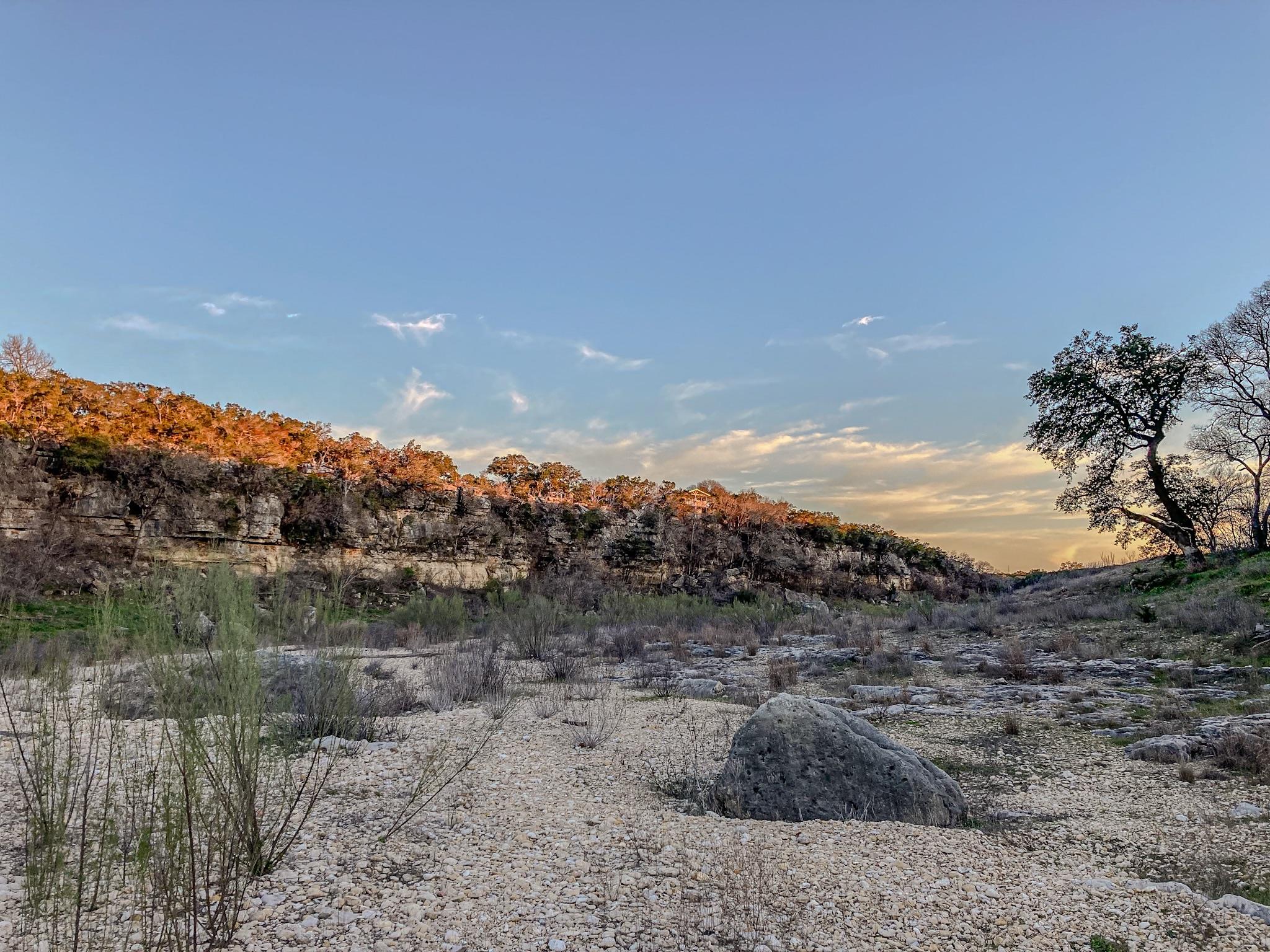 Rocky undeveloped land below a limestone cliff, with a large oak tree and golden light.