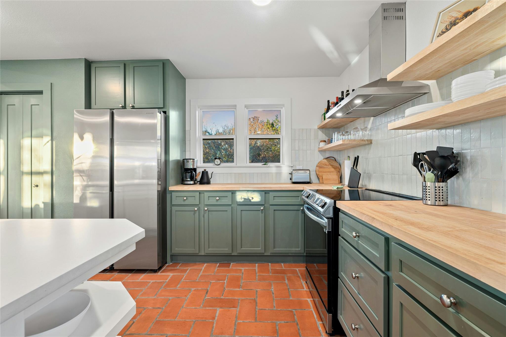 Kitchen with sage-green cabinets, butcher-block counters, open shelves, and terracotta tile flooring.