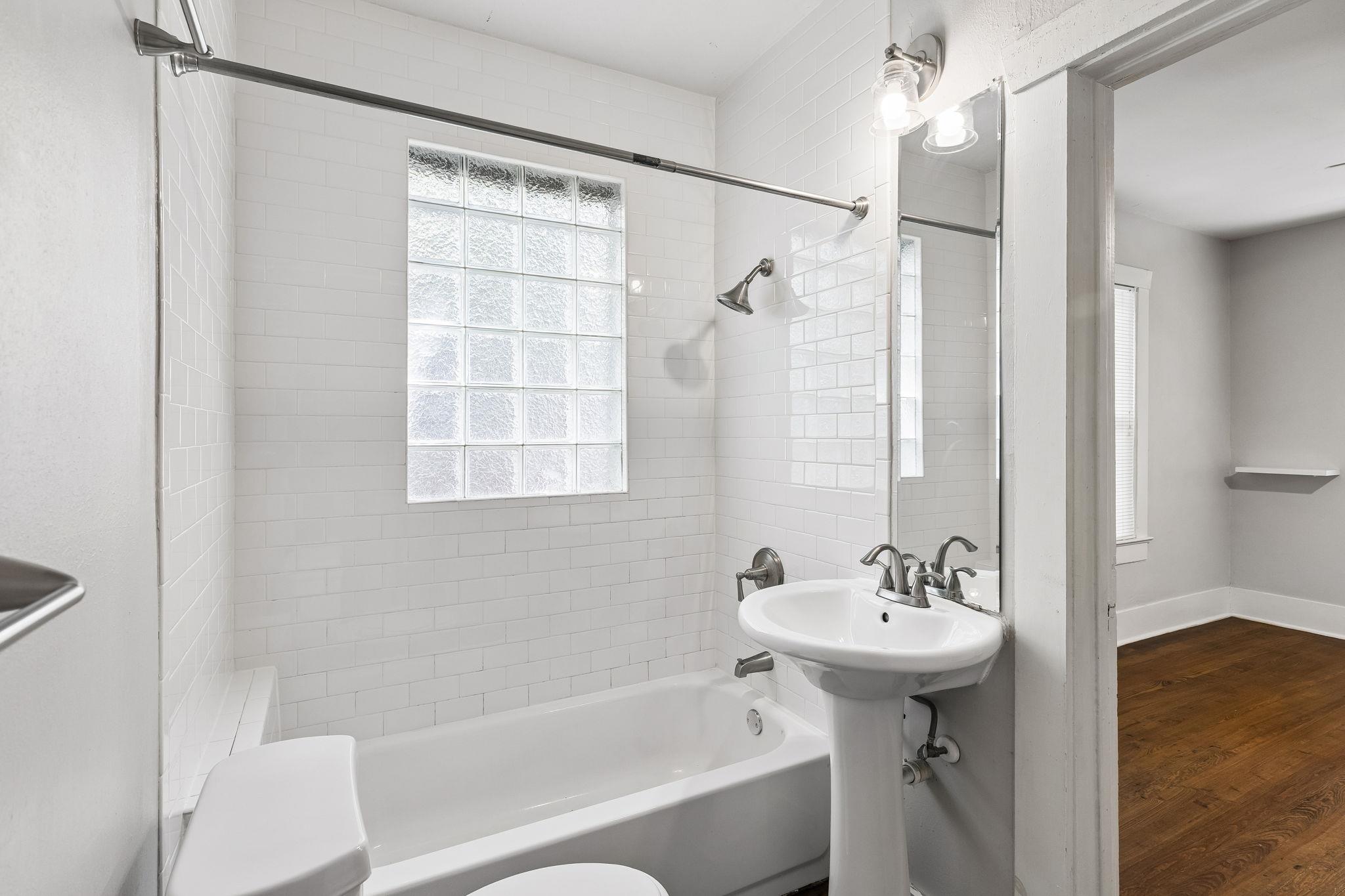 White tiled bathroom with a bathtub, glass-block window, and pedestal sink beside a hardwood-floored room.
