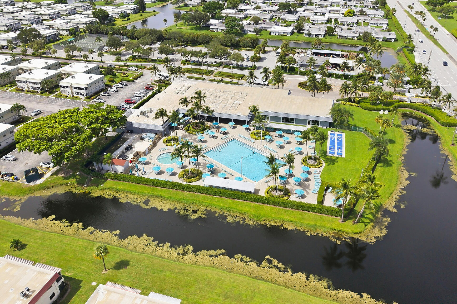 Aerial view of a community clubhouse and pool beside a canal, surrounded by palm trees and homes.
