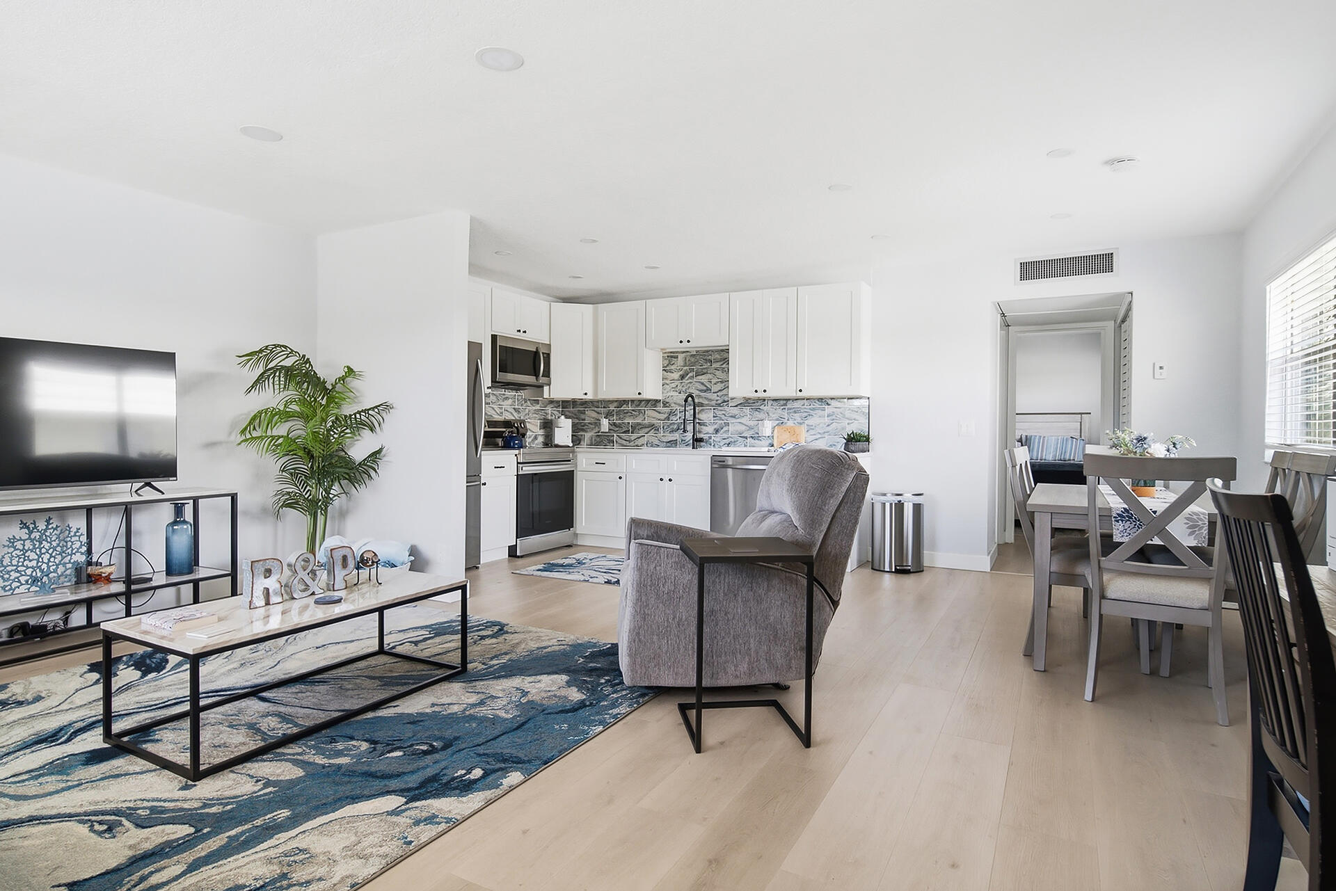 Open-plan living and dining area with white kitchen, blue patterned backsplash, and large blue-and-white rug.