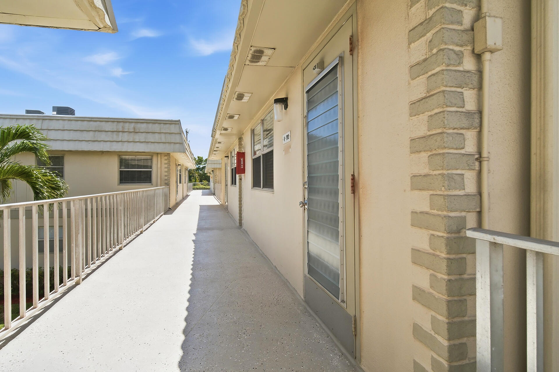 Residential building walkway with a unit entrance, white railings, and a long sunlit corridor.