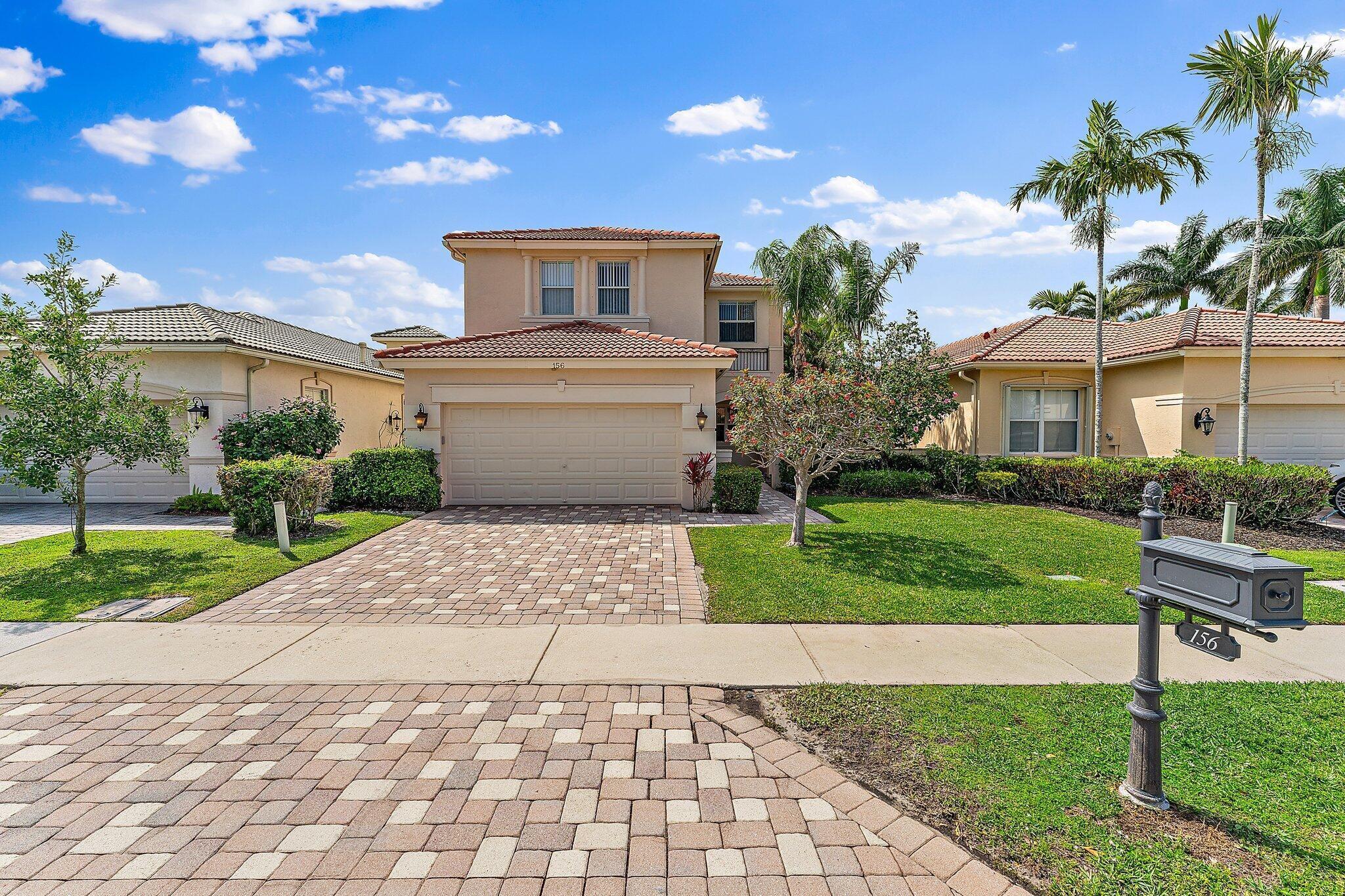 Two-story stucco home with terracotta roof, double garage, and paved driveway lined by palm trees.