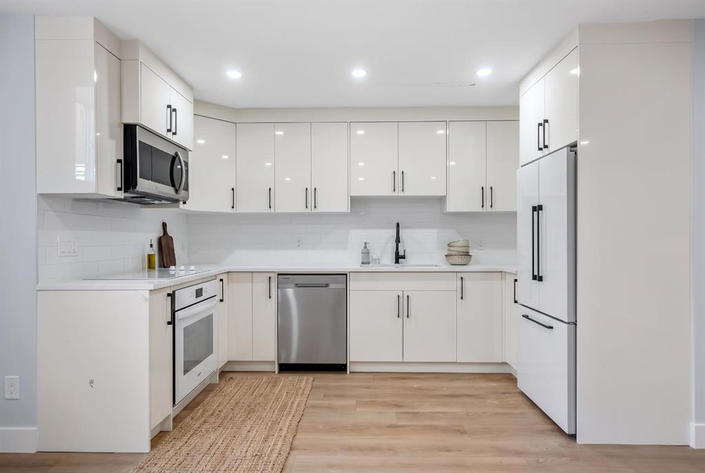 Modern white kitchen with stainless steel appliances, black faucet, and light wood flooring.