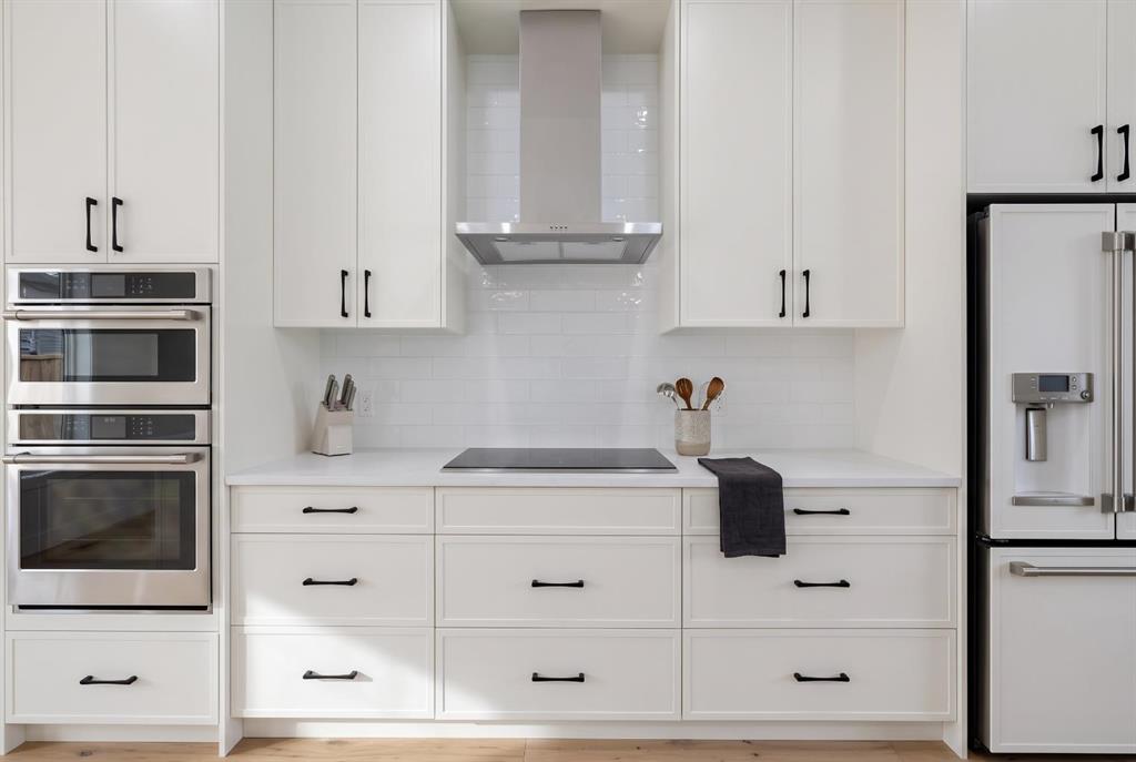 Modern white kitchen with built-in double ovens, induction cooktop, and stainless steel refrigerator.