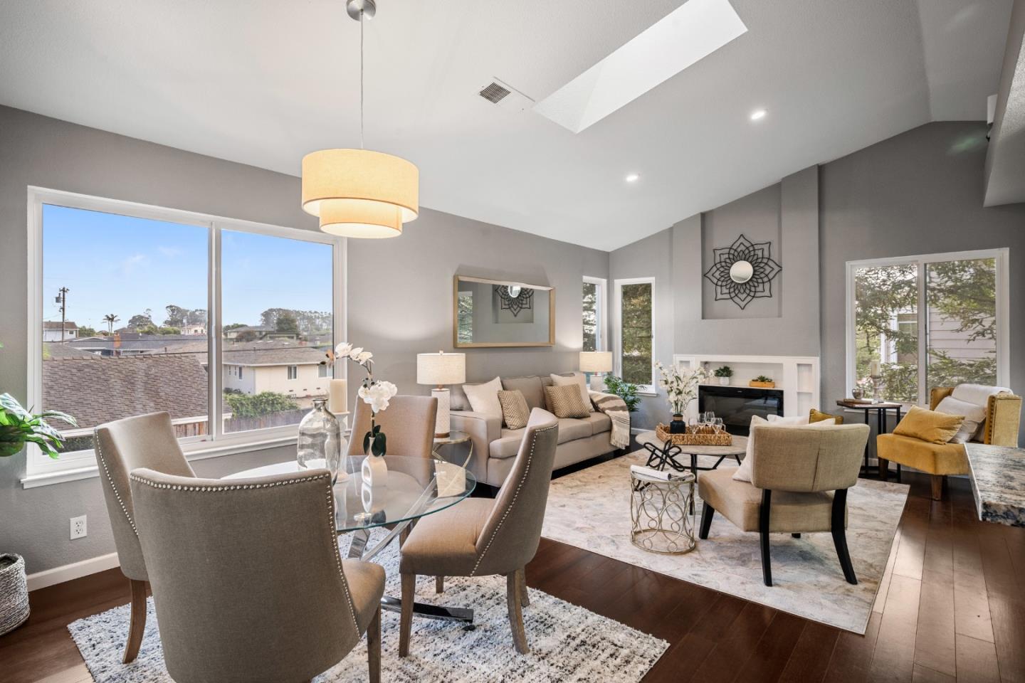 Open-plan living and dining room with a glass table, neutral furnishings, fireplace, and large windows.