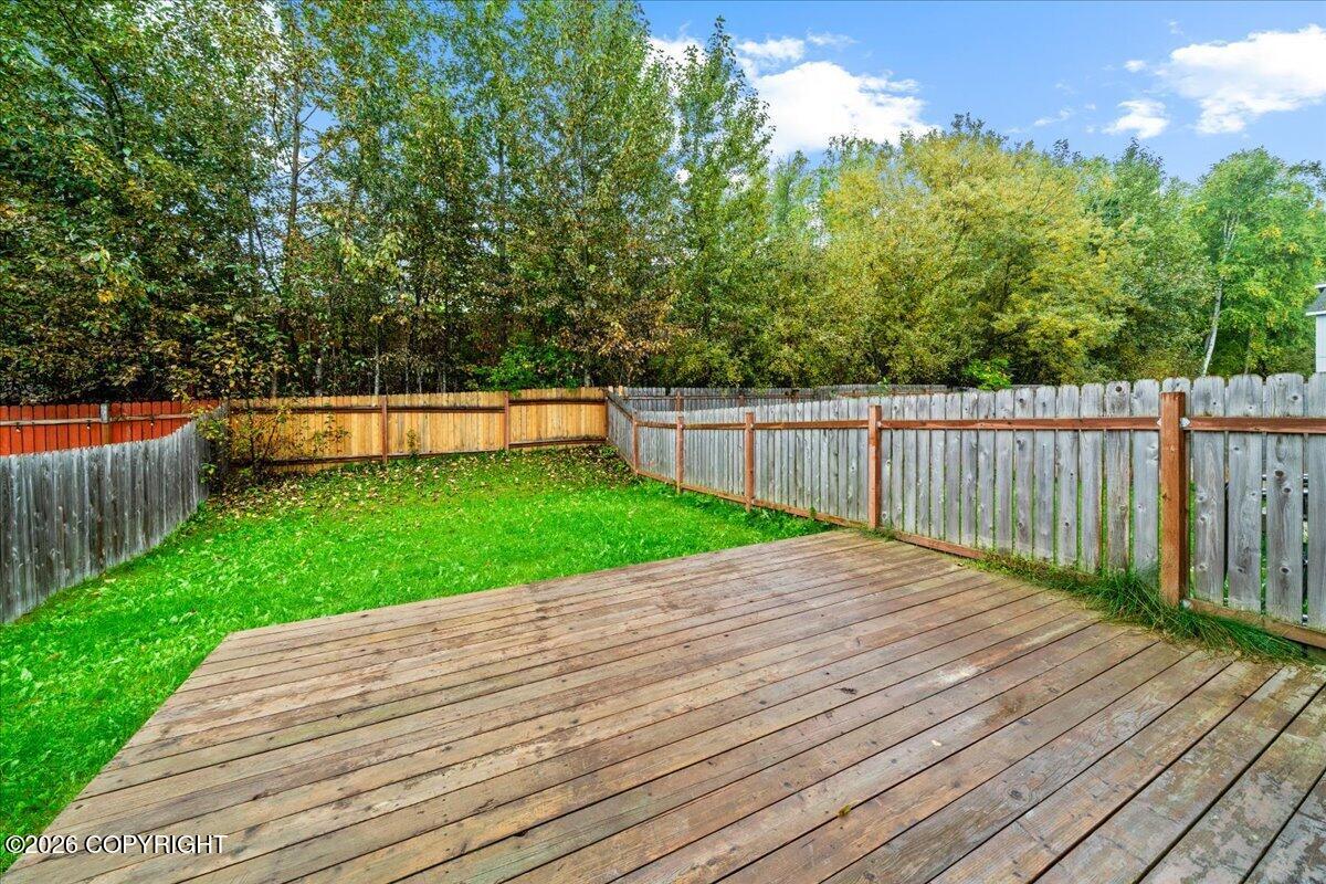 Fenced backyard with a large wooden deck, green lawn, and leafy trees.