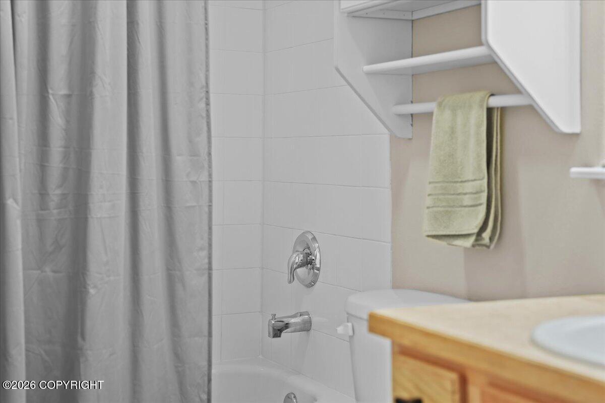 Bathroom tub and shower with white tile, gray curtain, chrome fixtures, and wood-tone vanity.
