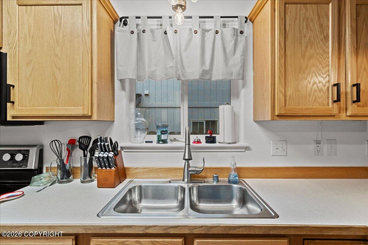 Kitchen sink with double stainless-steel basins, tall faucet, wood cabinets, and a curtained window.