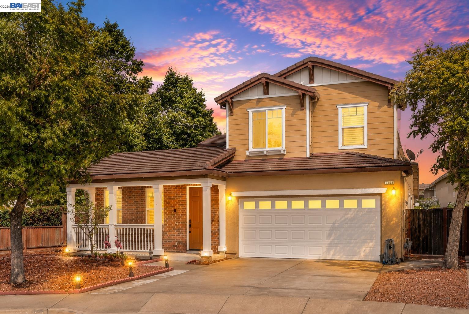 Two-story tan-and-brick home with a covered porch and attached garage beneath a vivid sunset.