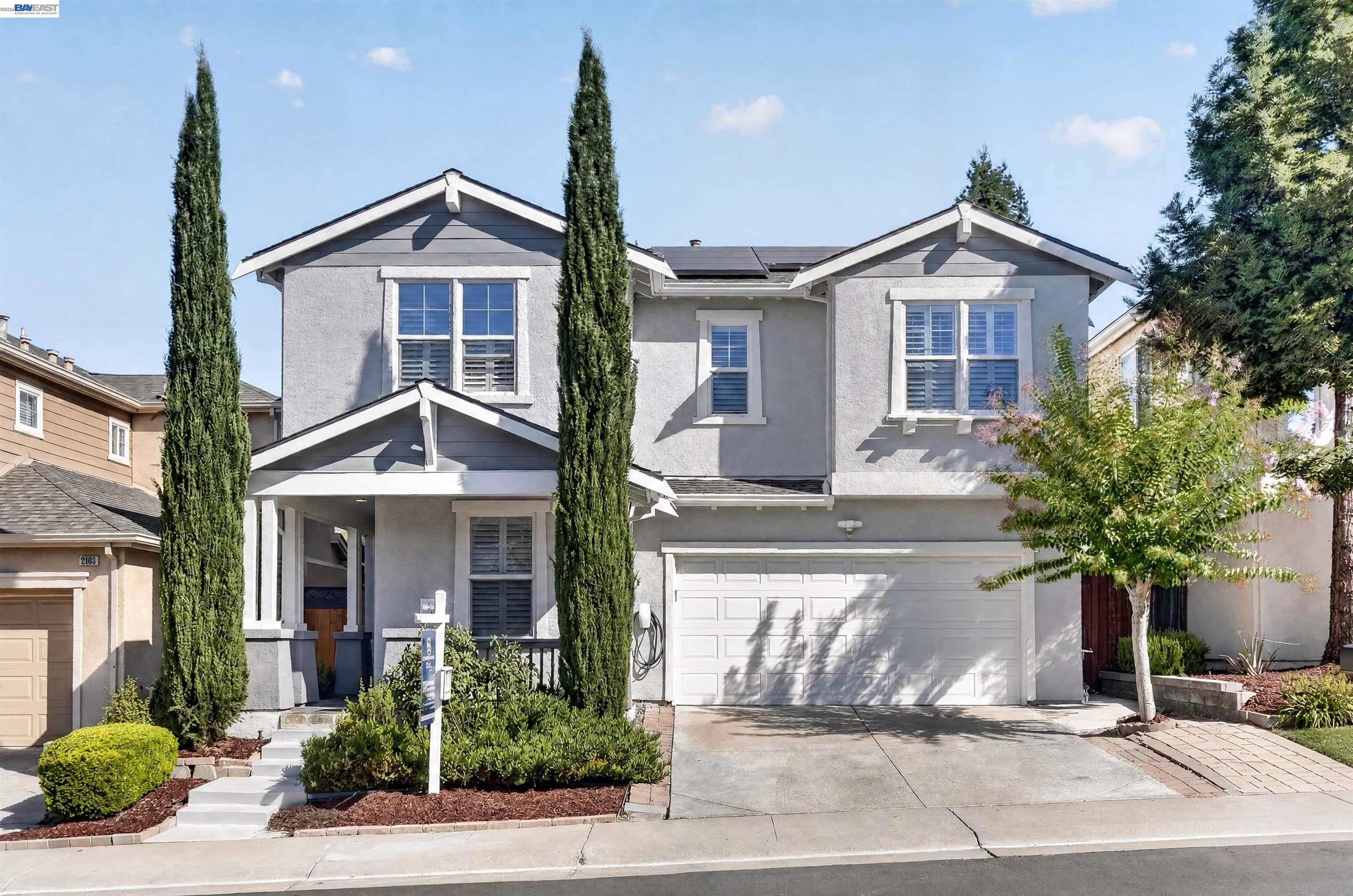 Two-story gray home with solar panels, a white garage door, and tall cypress trees.
