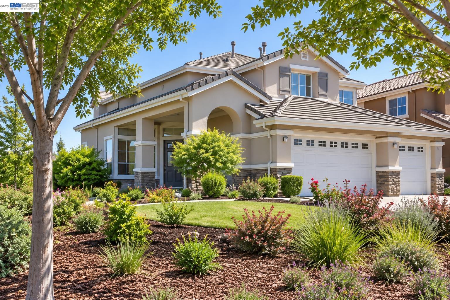 Two-story beige home with an arched entry, tiled roof, two-car garage, and landscaped front yard.