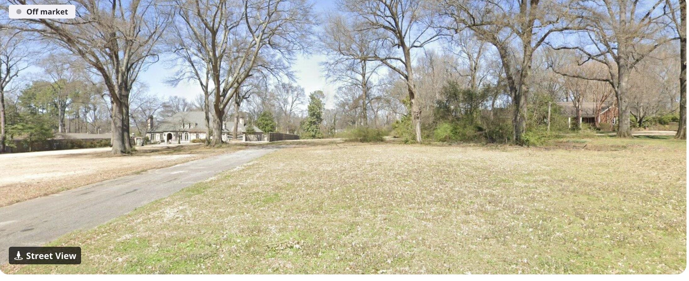 Residential lot with a broad lawn, mature leafless trees, and a paved driveway.