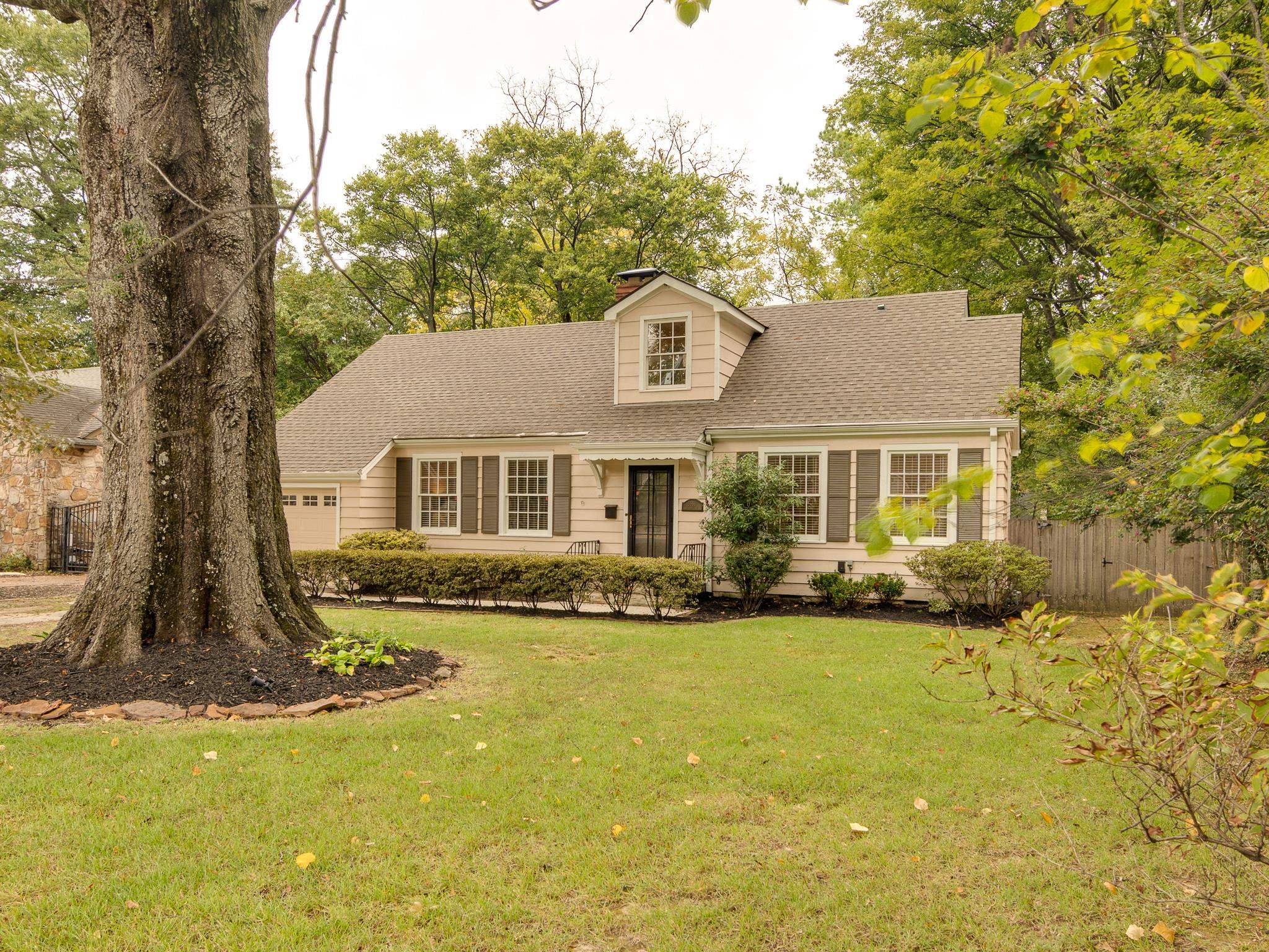 Beige house with a dormer and dark shutters, framed by a large tree and leafy yard.