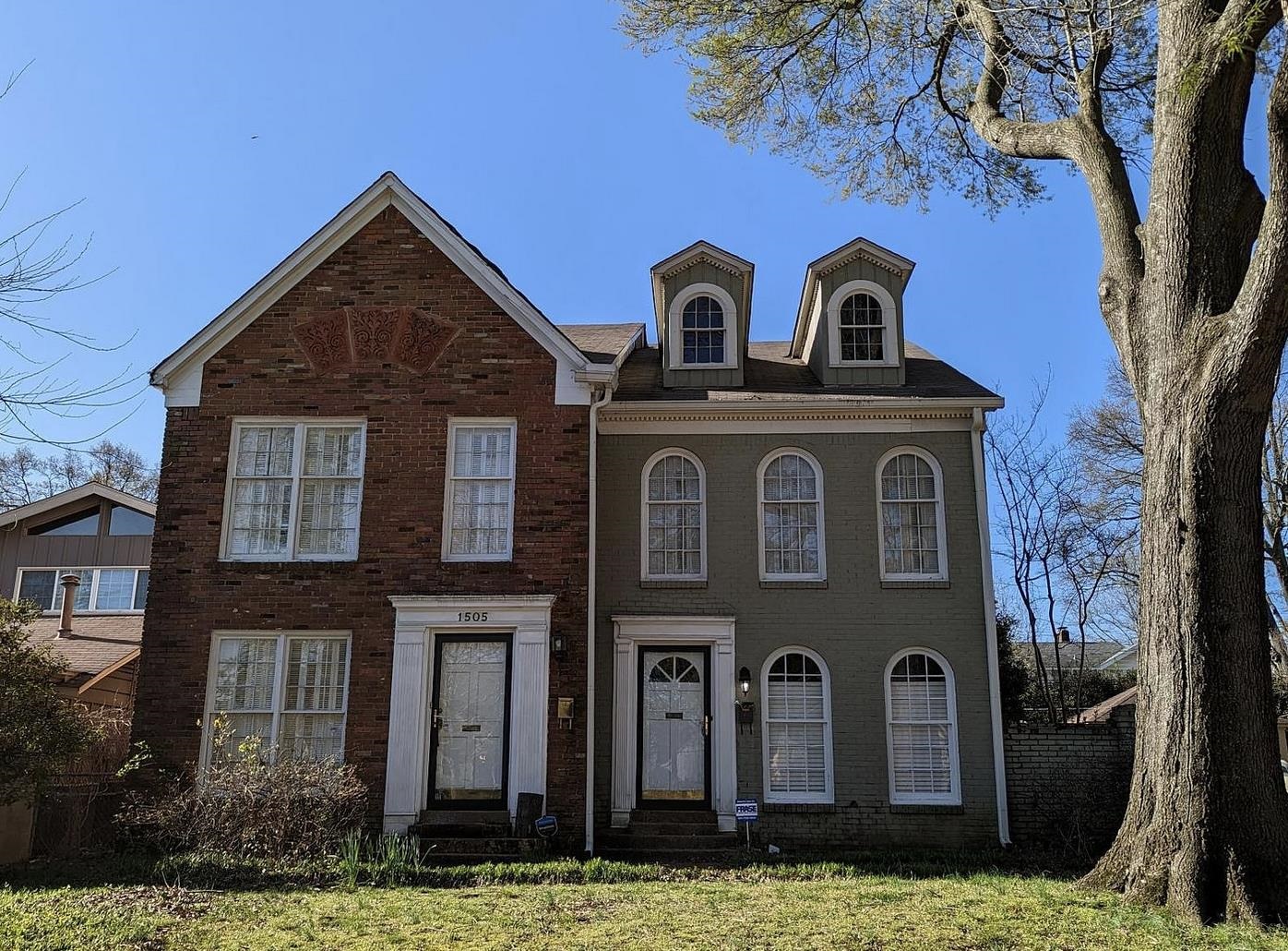 Attached brick and gray-green homes with arched windows, dormers, and a large tree.
