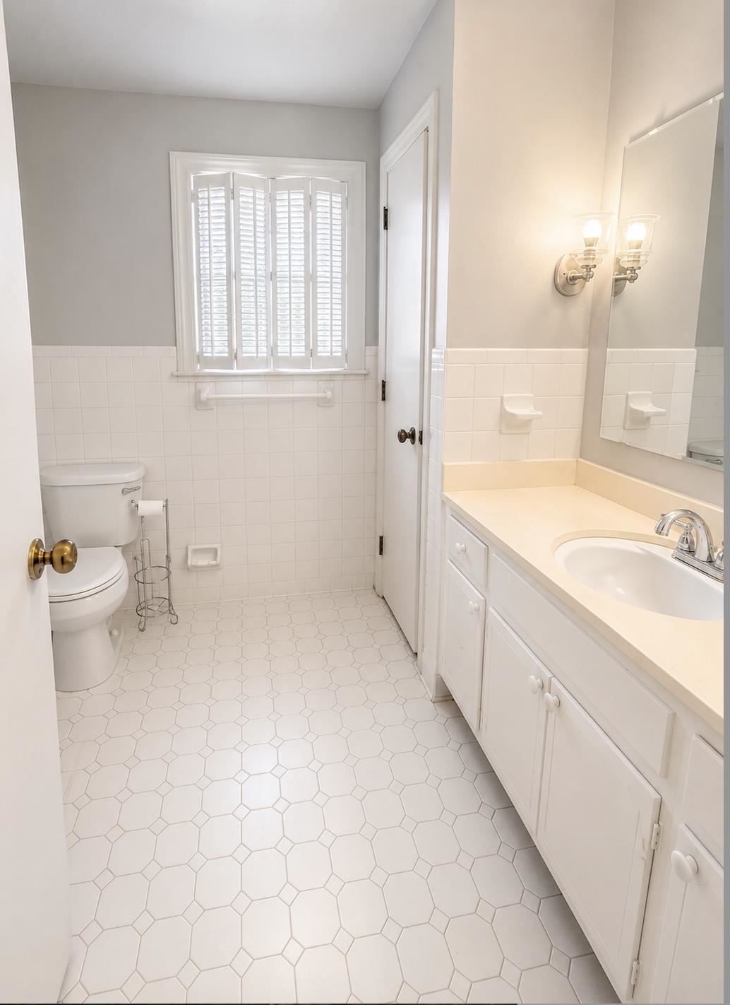 Bathroom with white hexagonal floor tiles, shuttered window, toilet, and long vanity with sink.