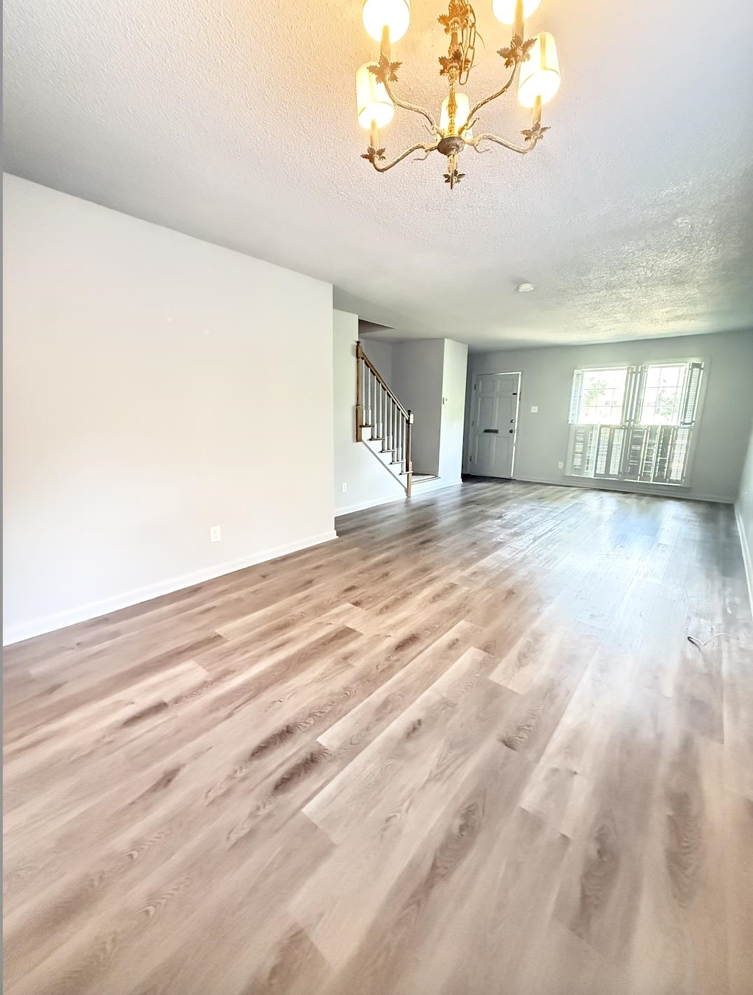 Empty living room with wood-look floors, a brass chandelier, staircase, and shuttered windows.