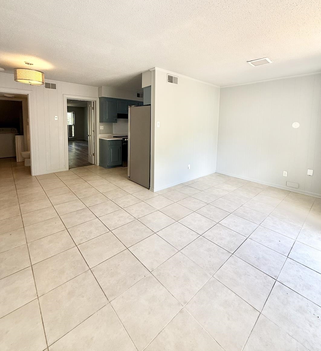 Unfurnished tiled room beside a compact kitchen with dark teal cabinets and a refrigerator.