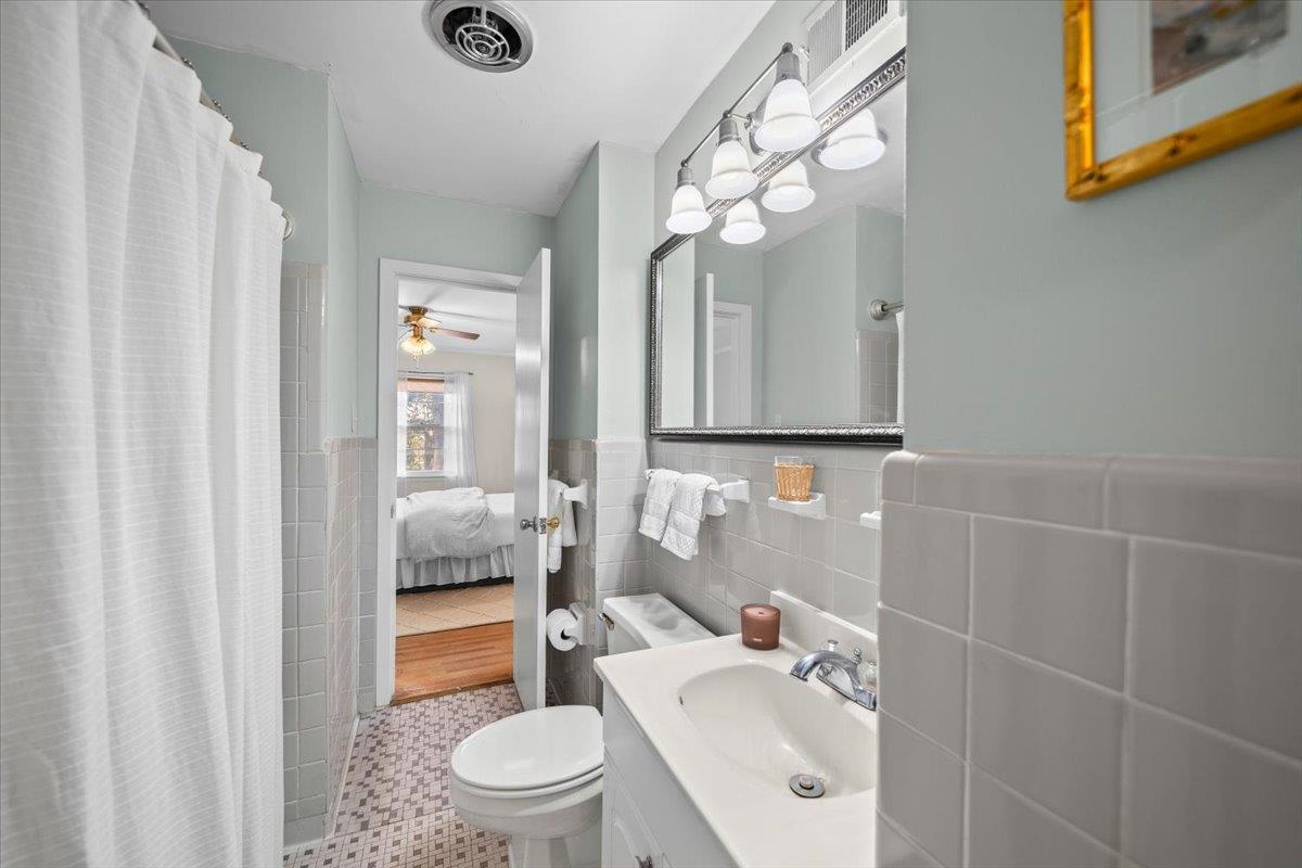 Bathroom with white tiled walls, a vanity and mirror, and a curtained shower beside an open bedroom doorway.