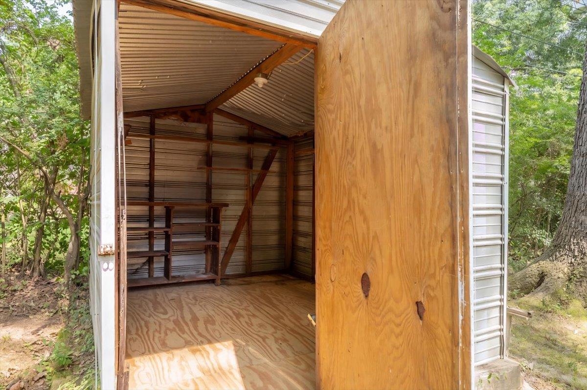 Open storage shed with plywood flooring, metal walls, and wooden shelving among trees.