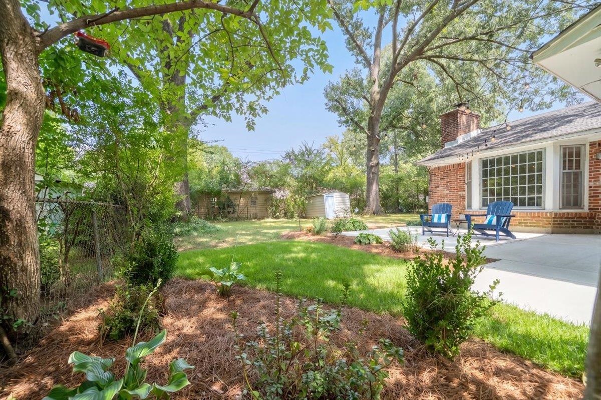 Shaded backyard with a lawn, mature trees, and a patio with blue chairs beside a brick house.