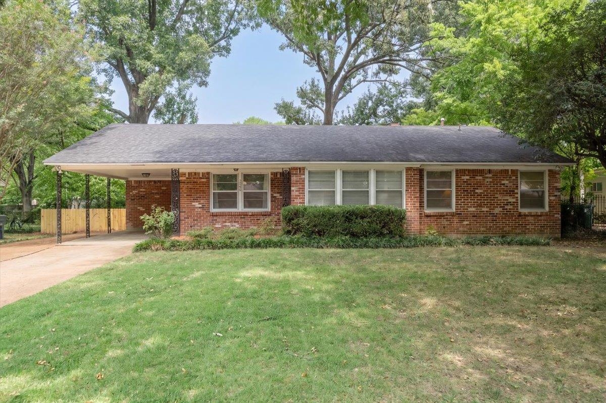 Single-story brick ranch house with a covered carport, broad lawn, and mature trees.