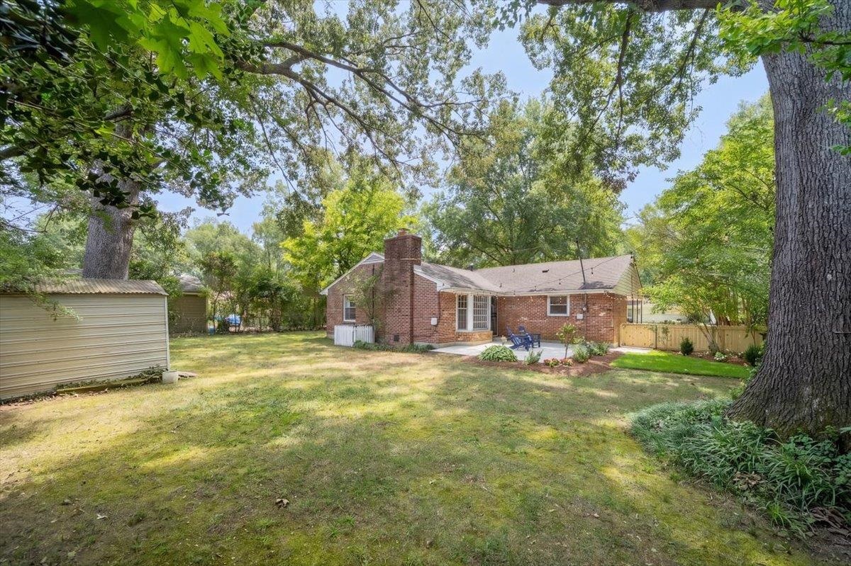 Brick home with a chimney and patio chairs beyond a spacious, tree-shaded backyard.