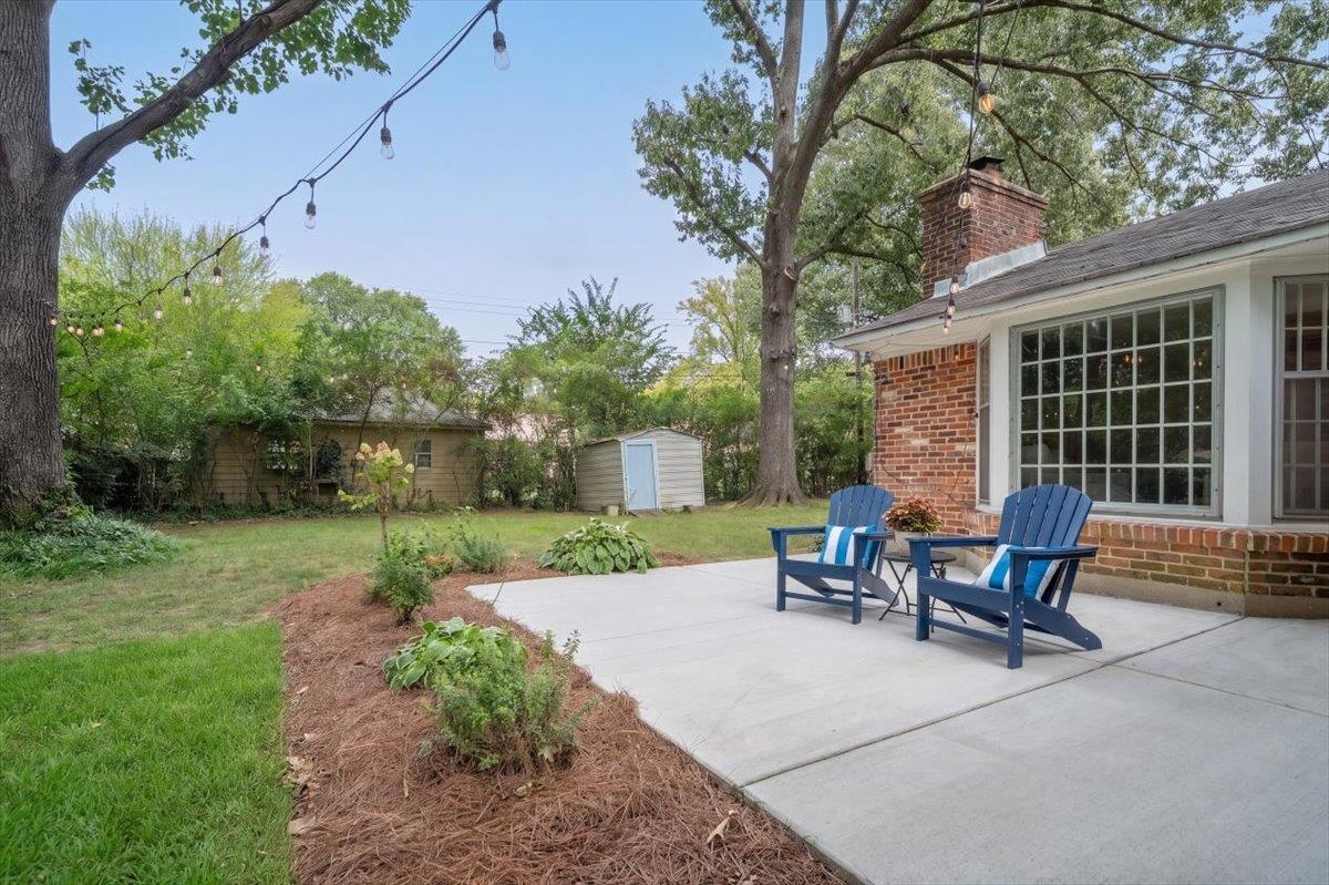 Backyard patio with blue Adirondack chairs beside a red-brick house, overlooking a lawn and mature trees.