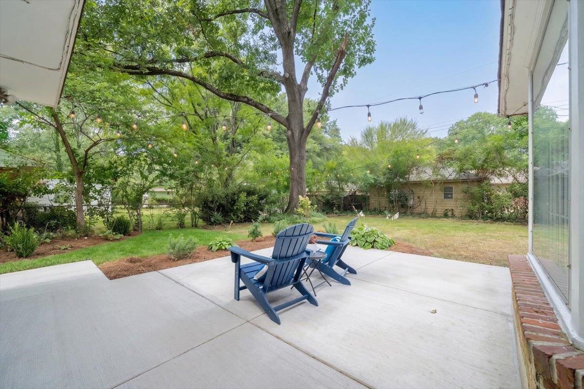 Concrete backyard patio with blue Adirondack chairs beneath mature trees and string lights.