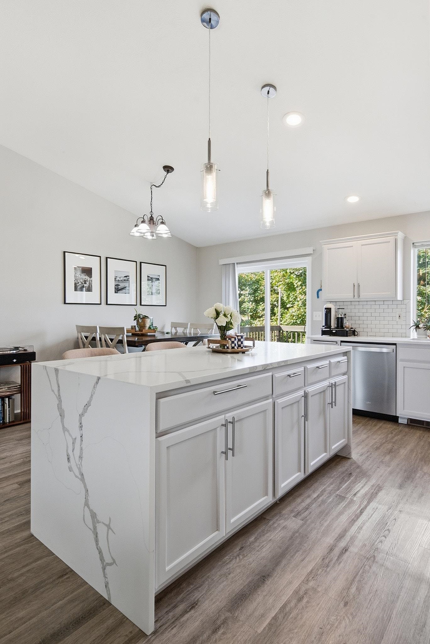 Bright kitchen with a large veined island, white cabinetry, pendant lights, and dining area by a glass door.