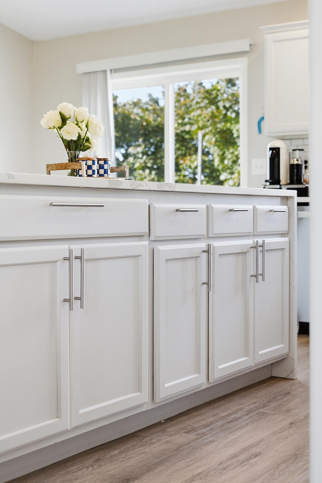 Bright kitchen with white Shaker cabinets, a long counter, and a large window overlooking leafy trees.