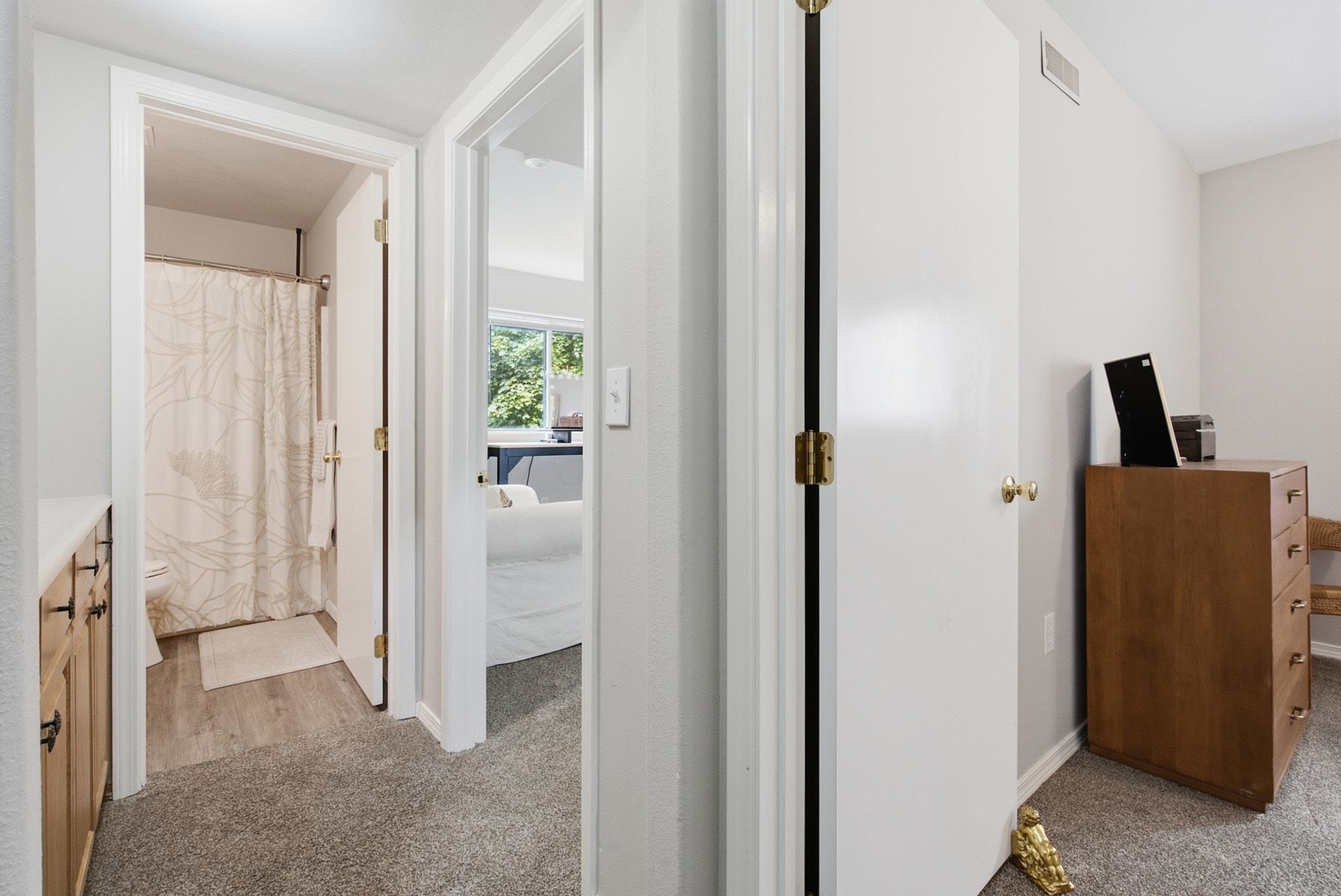 Carpeted hallway with open doors to a bathroom and bedroom, showing a patterned shower curtain and wooden dresser.