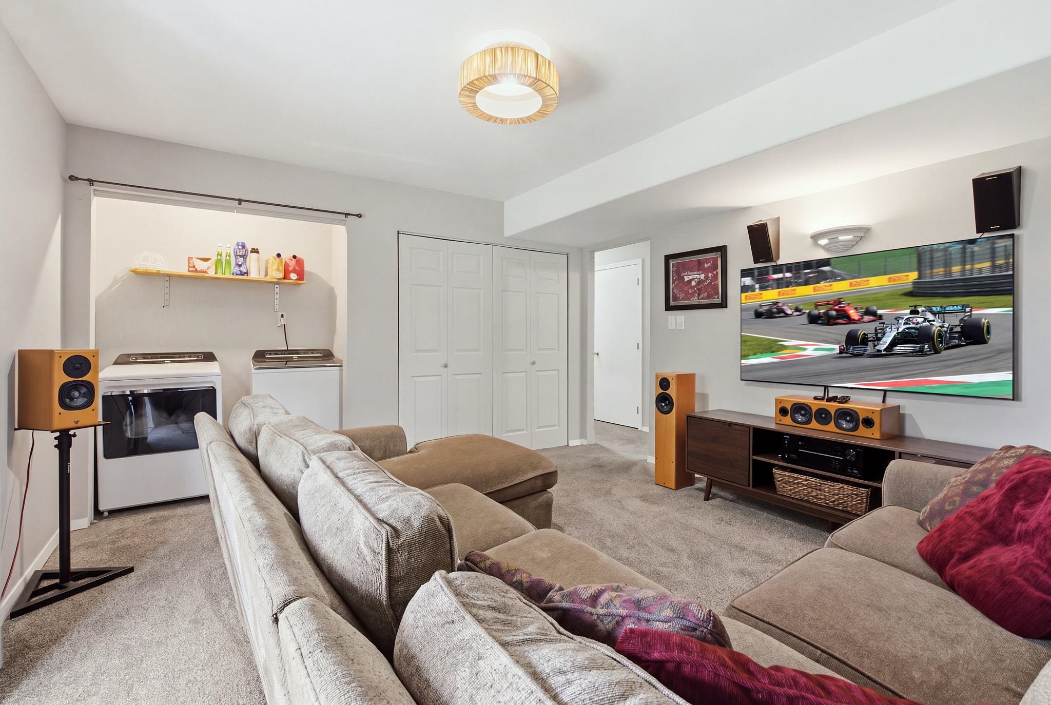 Furnished family room with a large sectional, race-car television, surround-sound speakers, and laundry appliances.