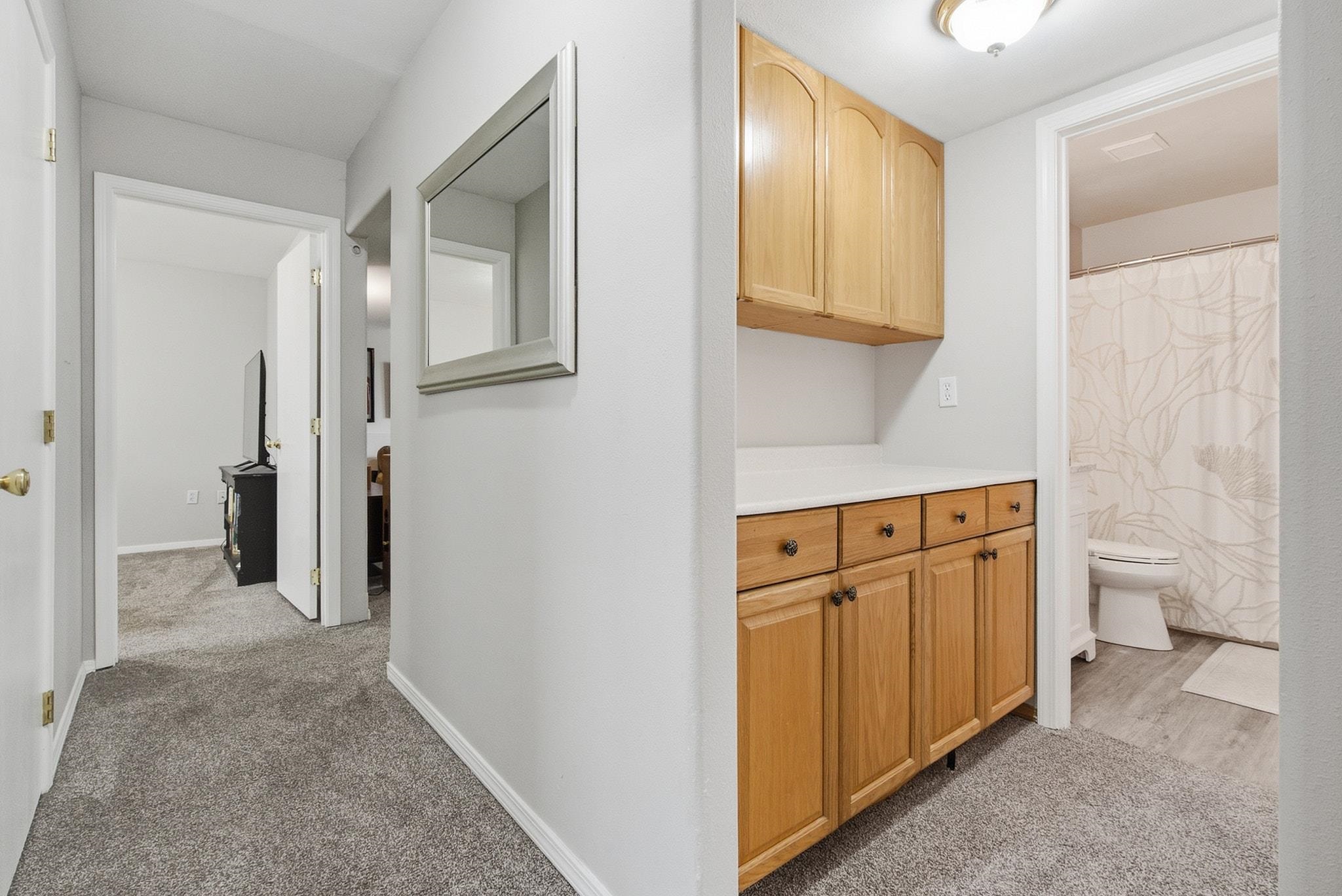 Bathroom vanity with honey-colored cabinets beside a doorway to a toilet and patterned shower curtain.