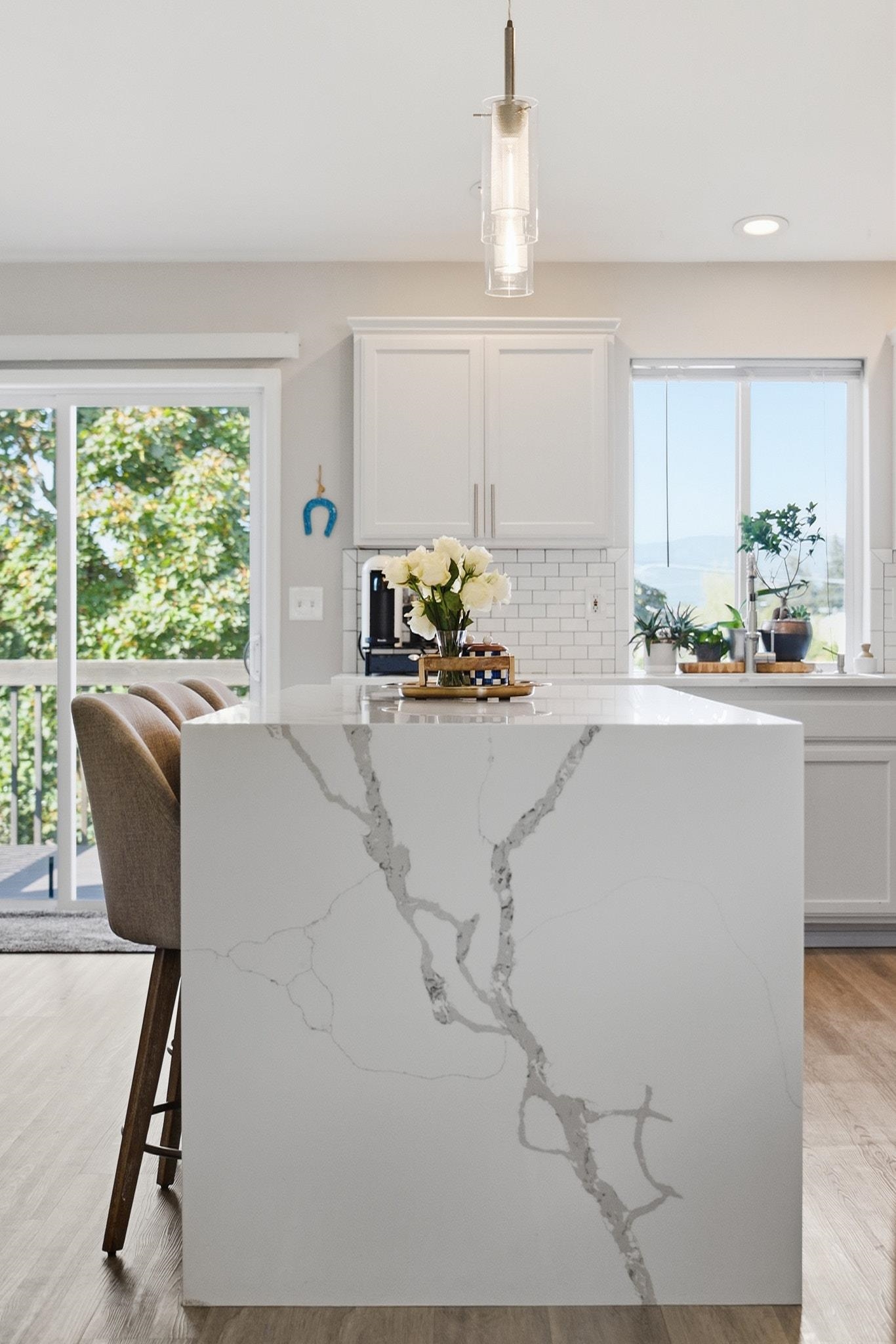 Kitchen with a large veined white island, white cabinetry, and windows overlooking greenery.