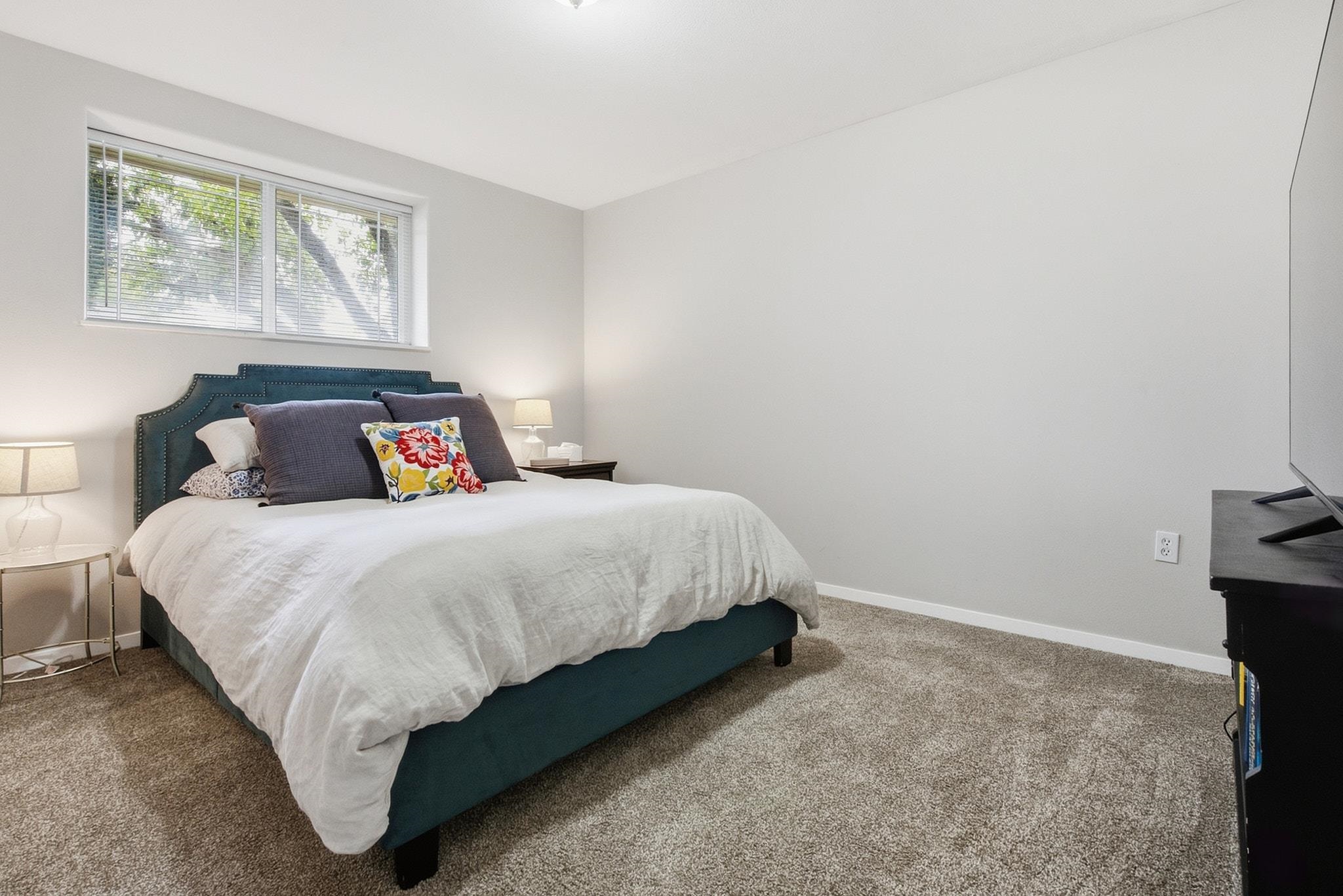 Staged bedroom with a teal bed, white bedding, beige carpet, and a wide window with blinds.
