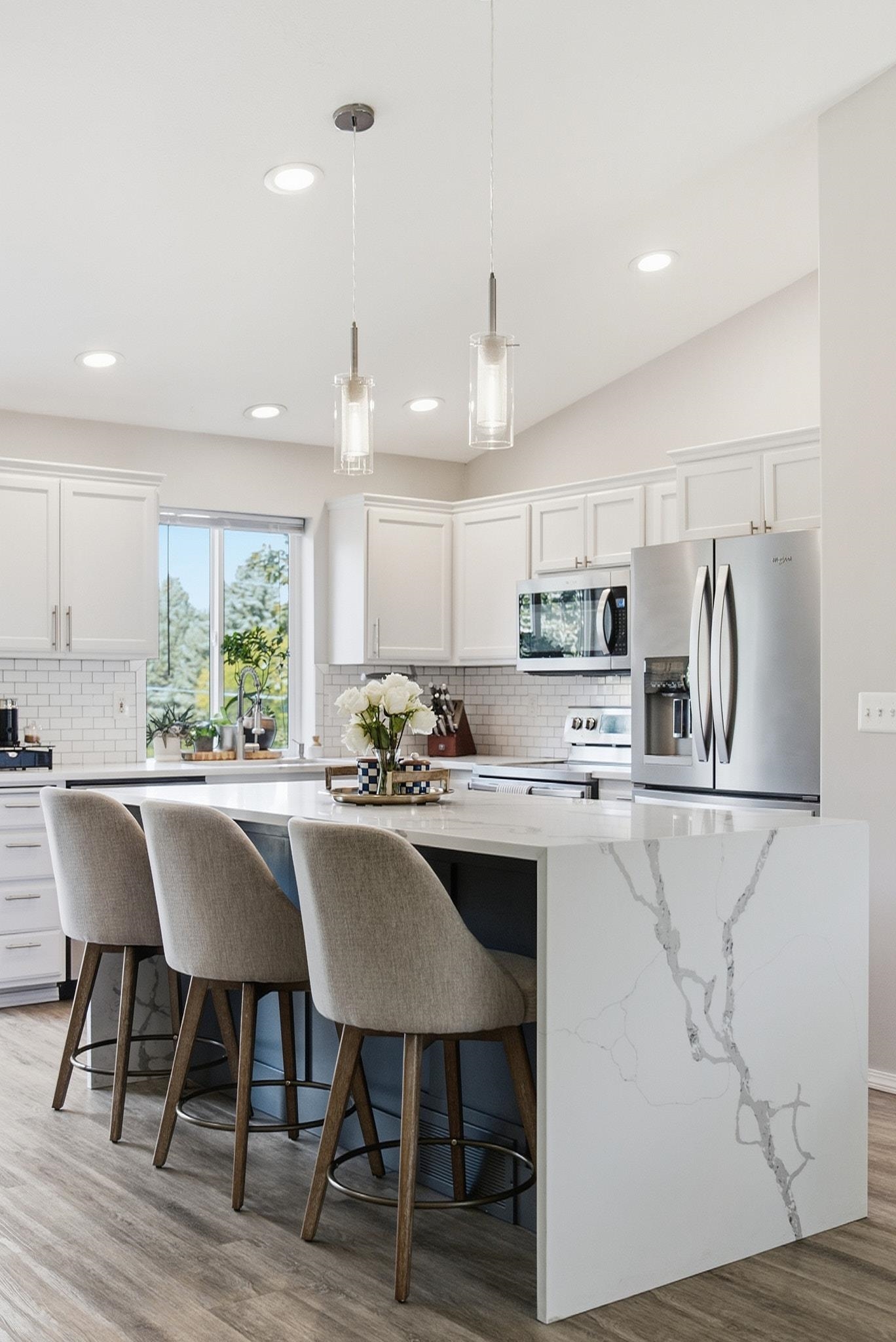Bright kitchen with a marble-look waterfall island, upholstered stools, white cabinets, and stainless steel appliances.